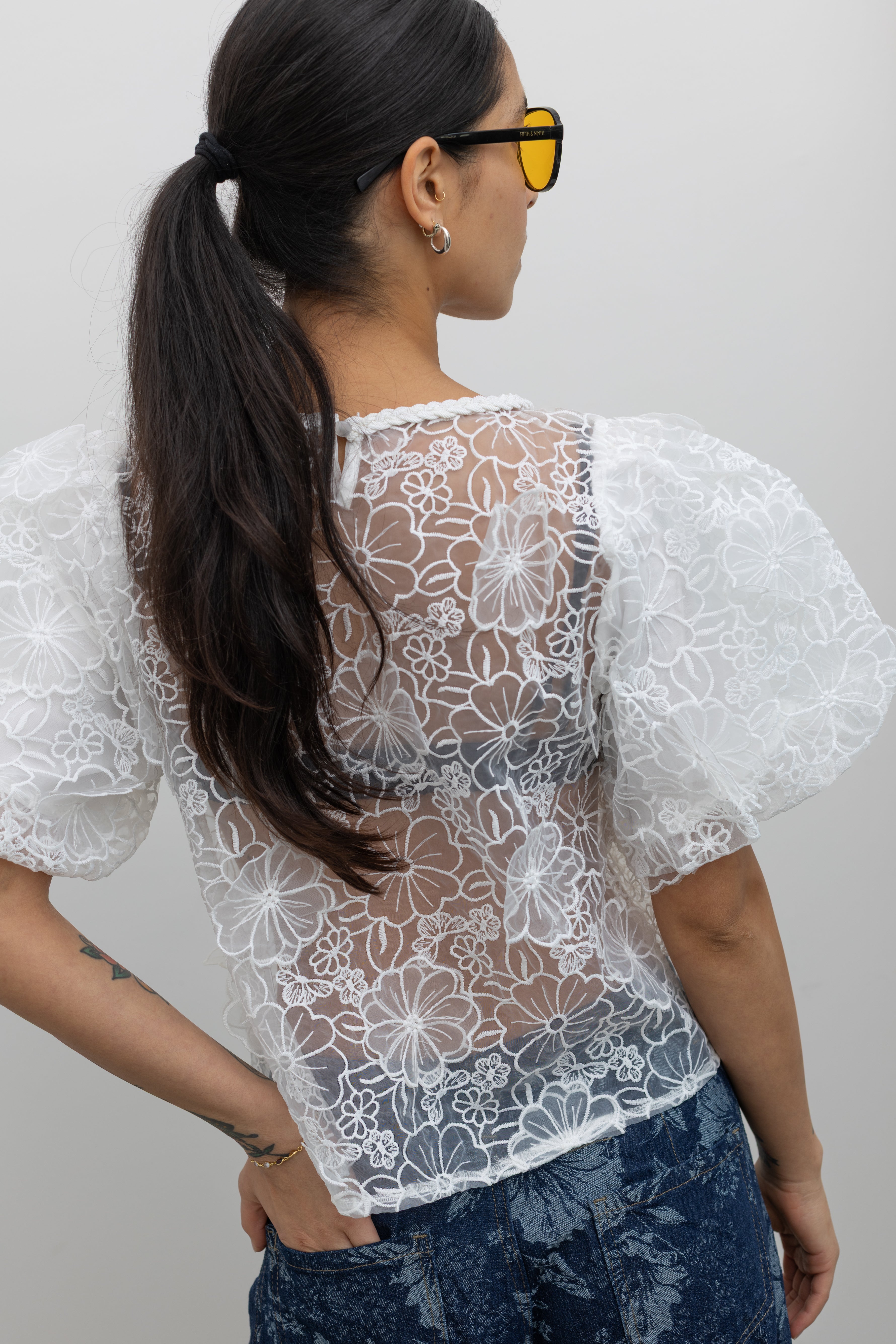 Back view of a woman posing wearing Spring fashion in a Collective Ivory Floral Embroidered Billow Sleeve Organza Blouse featuring a semi-sheer front paired with floral printed dark wash denim jeans at an Alas Eius store. 