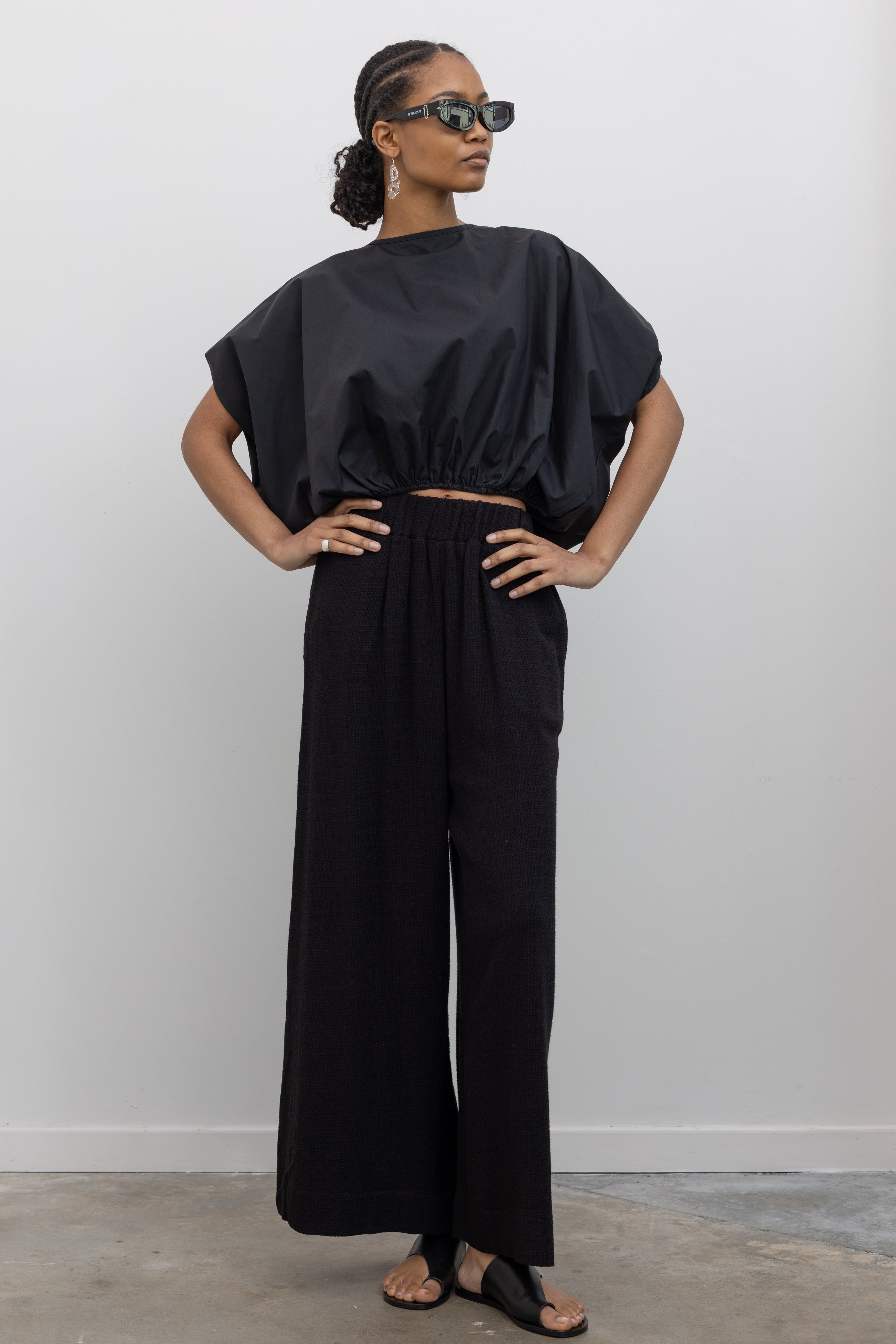 Full length front view of a woman posing wearing contemporary spring fashion in the Collective brand black linen wide leg pull on pants with a black balloon hem top and ATP black leather cutout sandals at an Alas Eius store. 