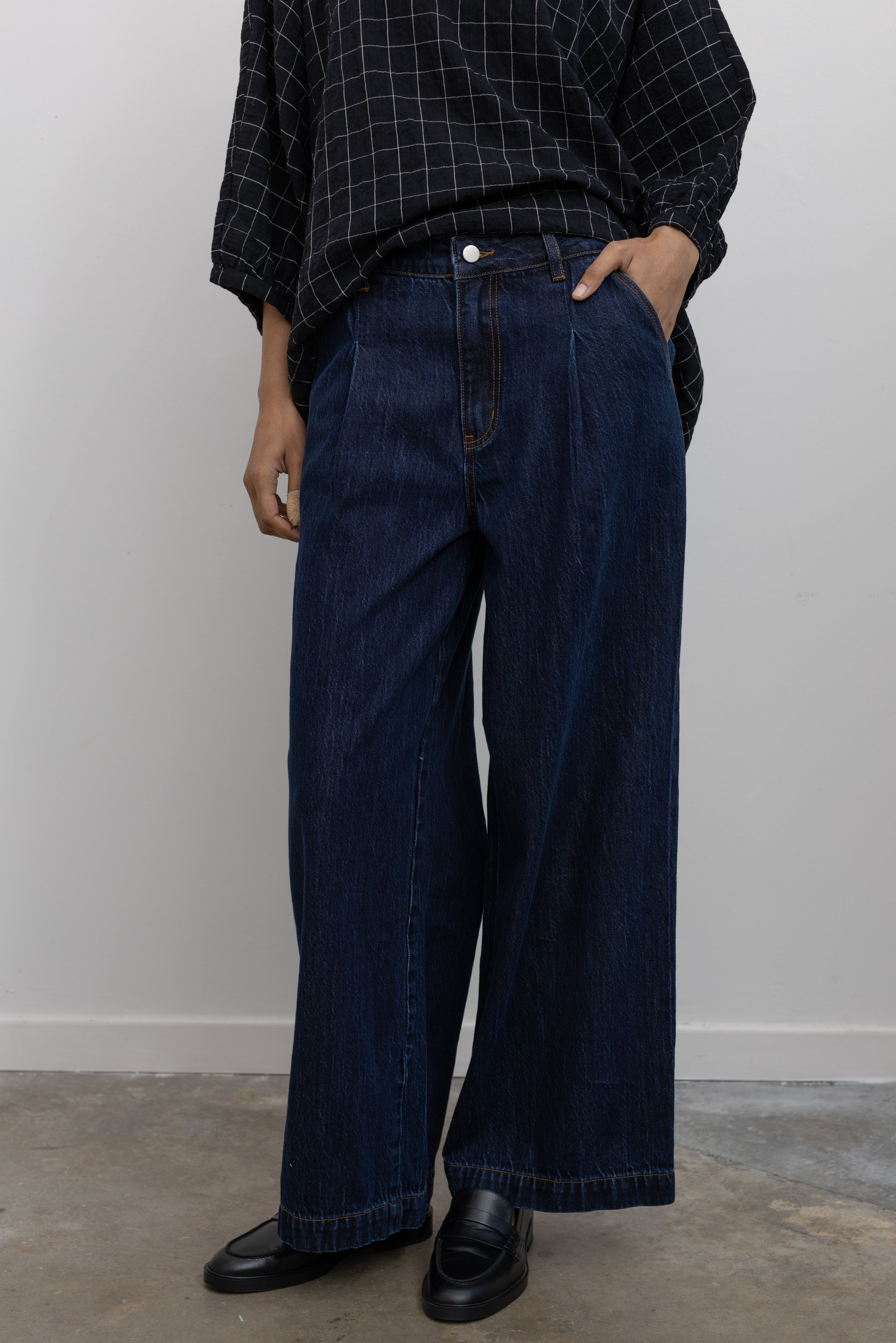 Close-up product view of a woman standing wearing a classic wardrobe staple in a pair of Collective indigo dark wash denim wide leg pleated jeans with a black top and black leather loafers at Alas Eius.