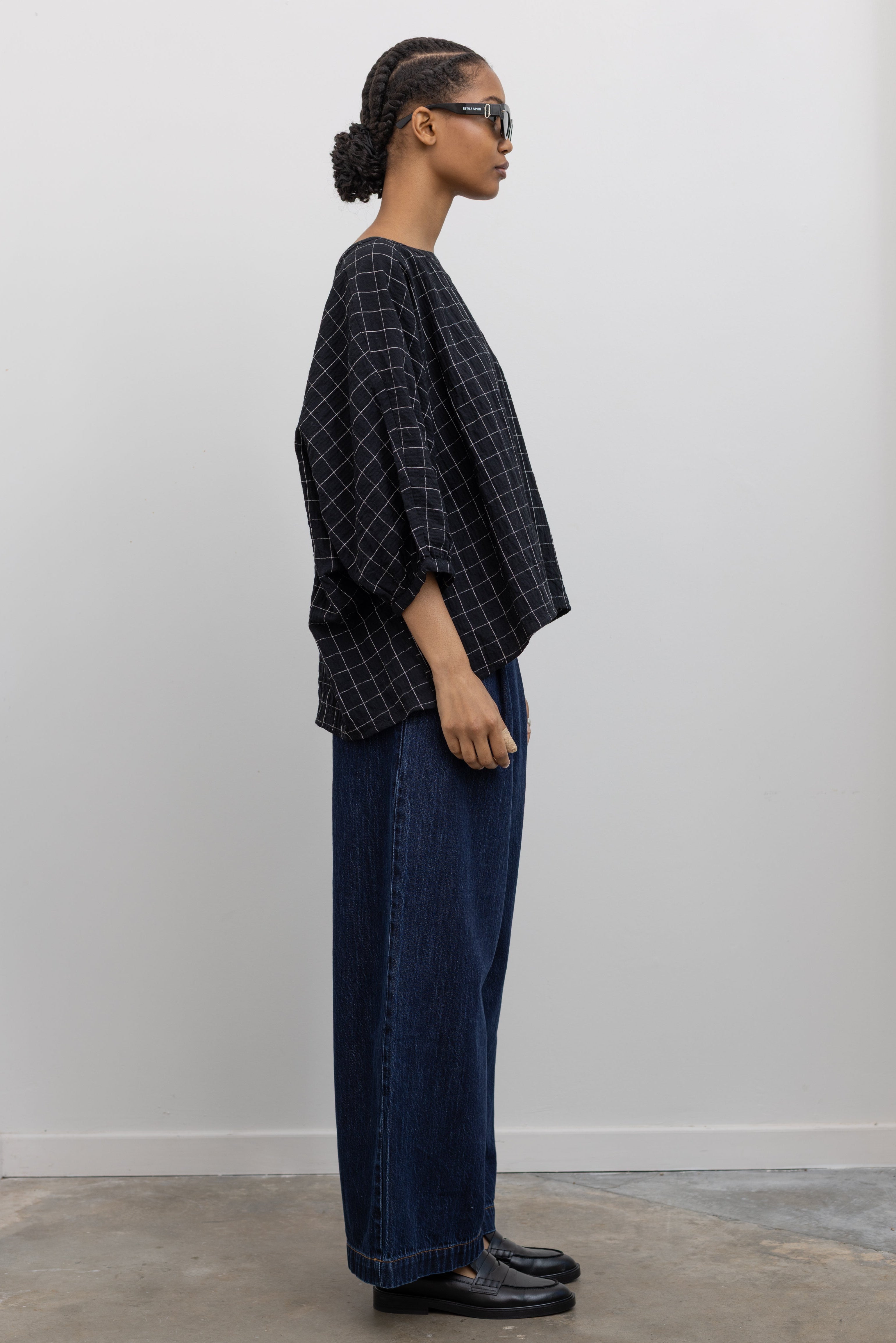 Side view of a woman standing wearing a classic wardrobe staple in a pair of Collective indigo denim wide leg pleated jeans with a black top and black leather loafers at Alas Eius.