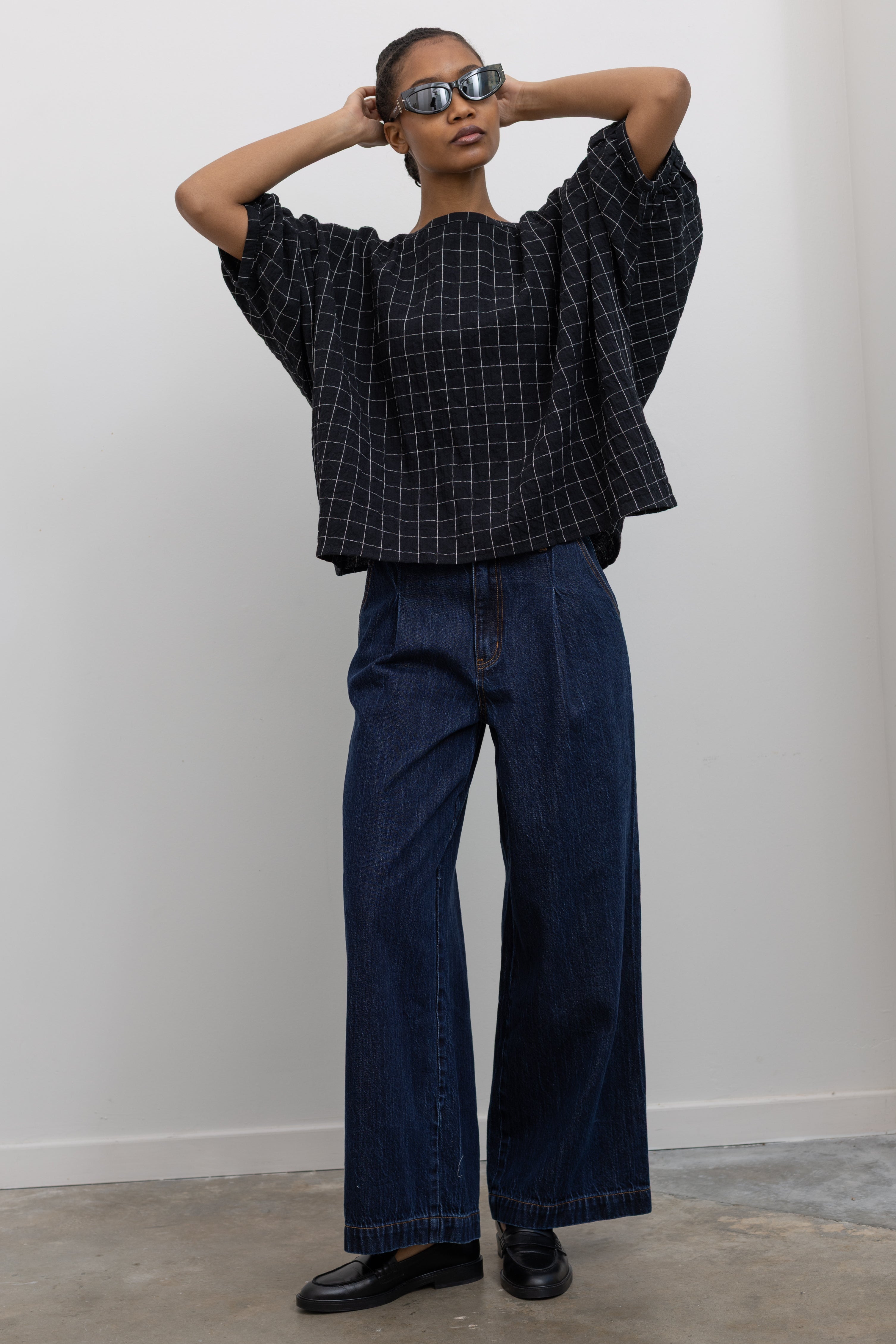 Woman posing with hands behind her head wearing a classic wardrobe staple in a pair of Collective indigo denim wide leg pleated jeans with a black top and black leather loafers at Alas Eius.