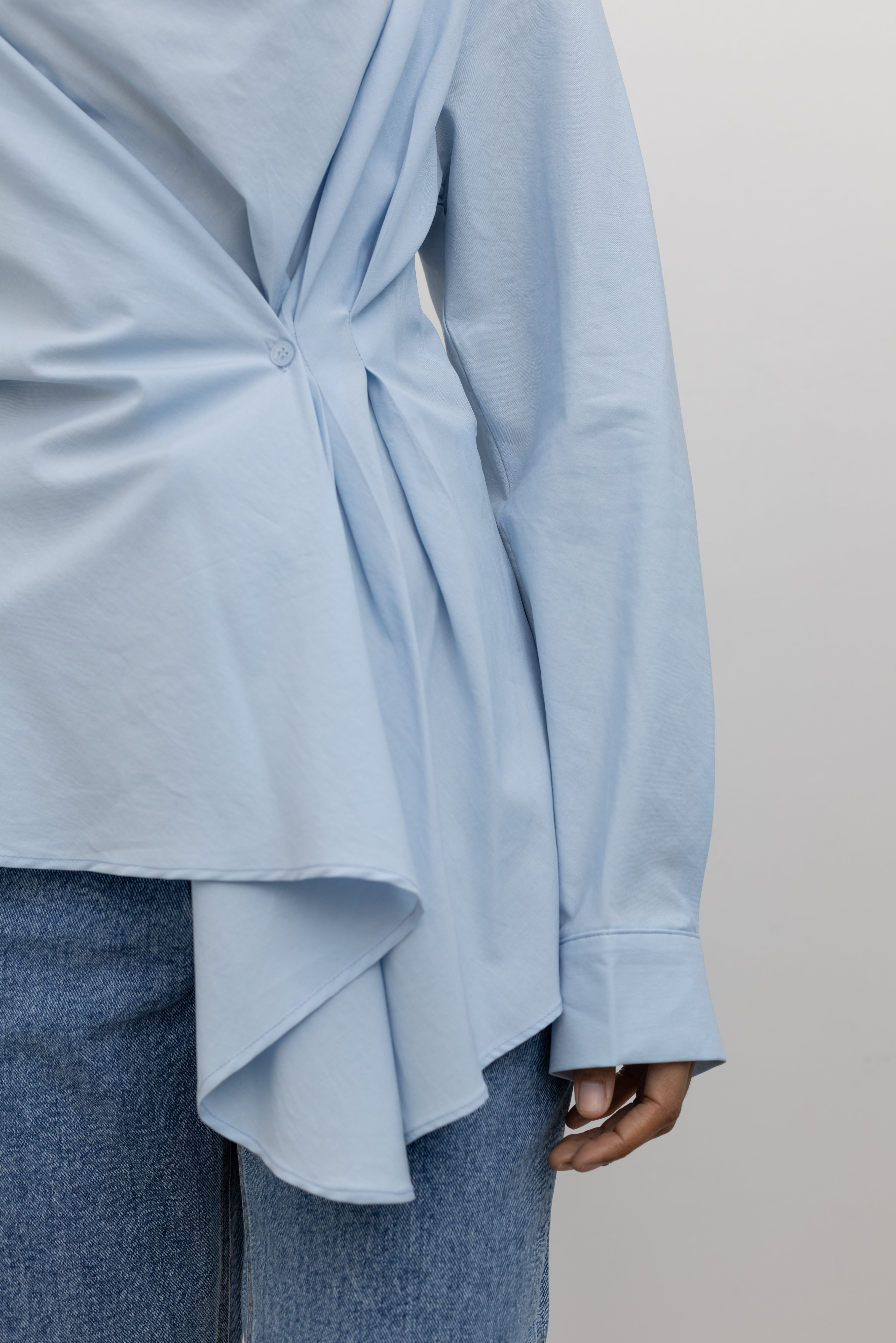 Close-up detail of gathered side and button detail at natural waistline on a Woman wearing a Collective label sky blue cotton long sleeve top featuring a fitted natural waistline with a side draped motif creating an asymmetrical hem and cowl neckline paired with light wash denim jeans at Alas Eius. 