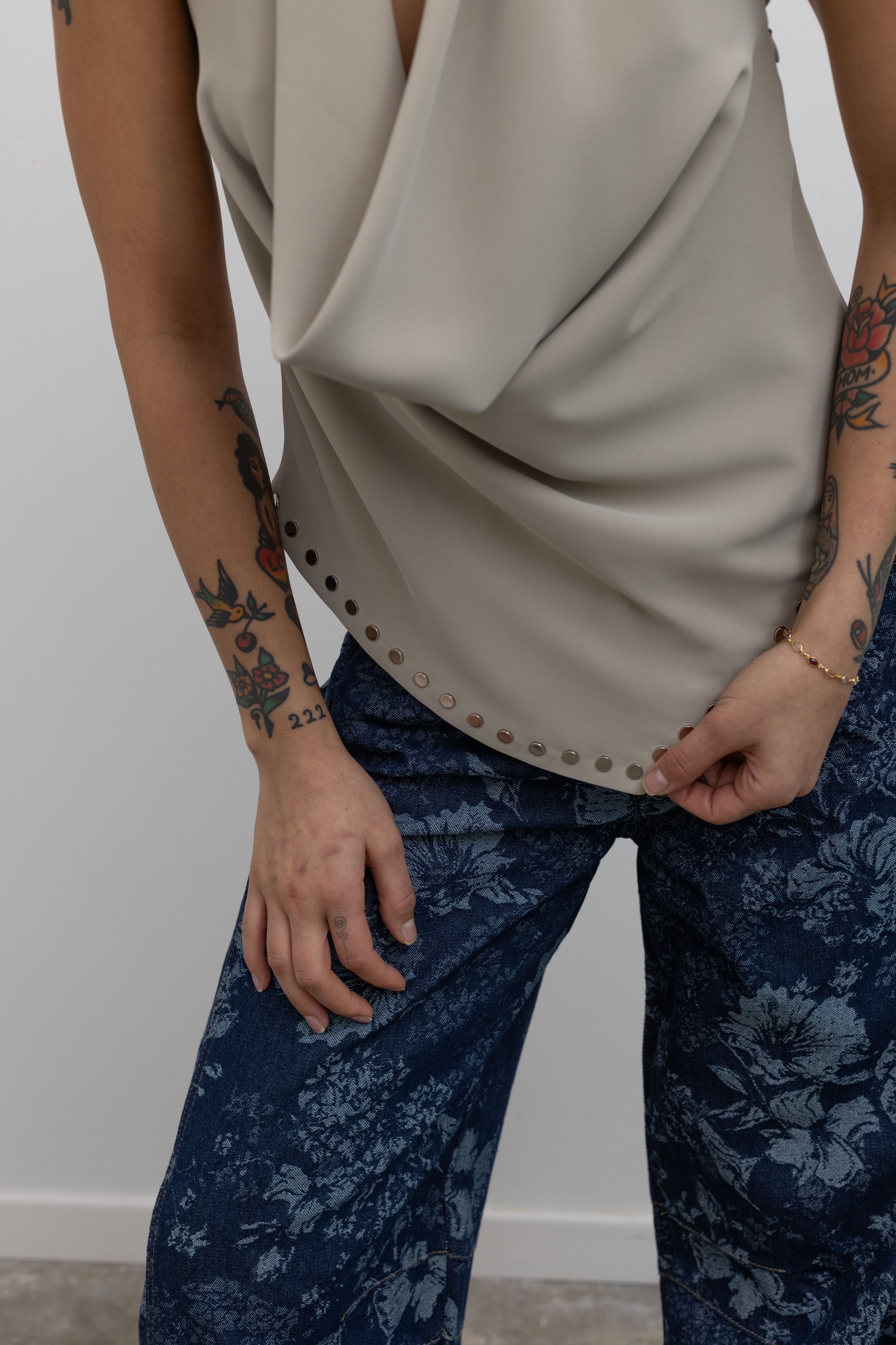 Close-up view of a woman wearing an edgy yet chic top in a Collective brand beige halter neck top designed with a low cowl drape, a v-cut hem that is lined with silver stud hardware, a side zipper closure, and exposed back at an Alas Eius Store.