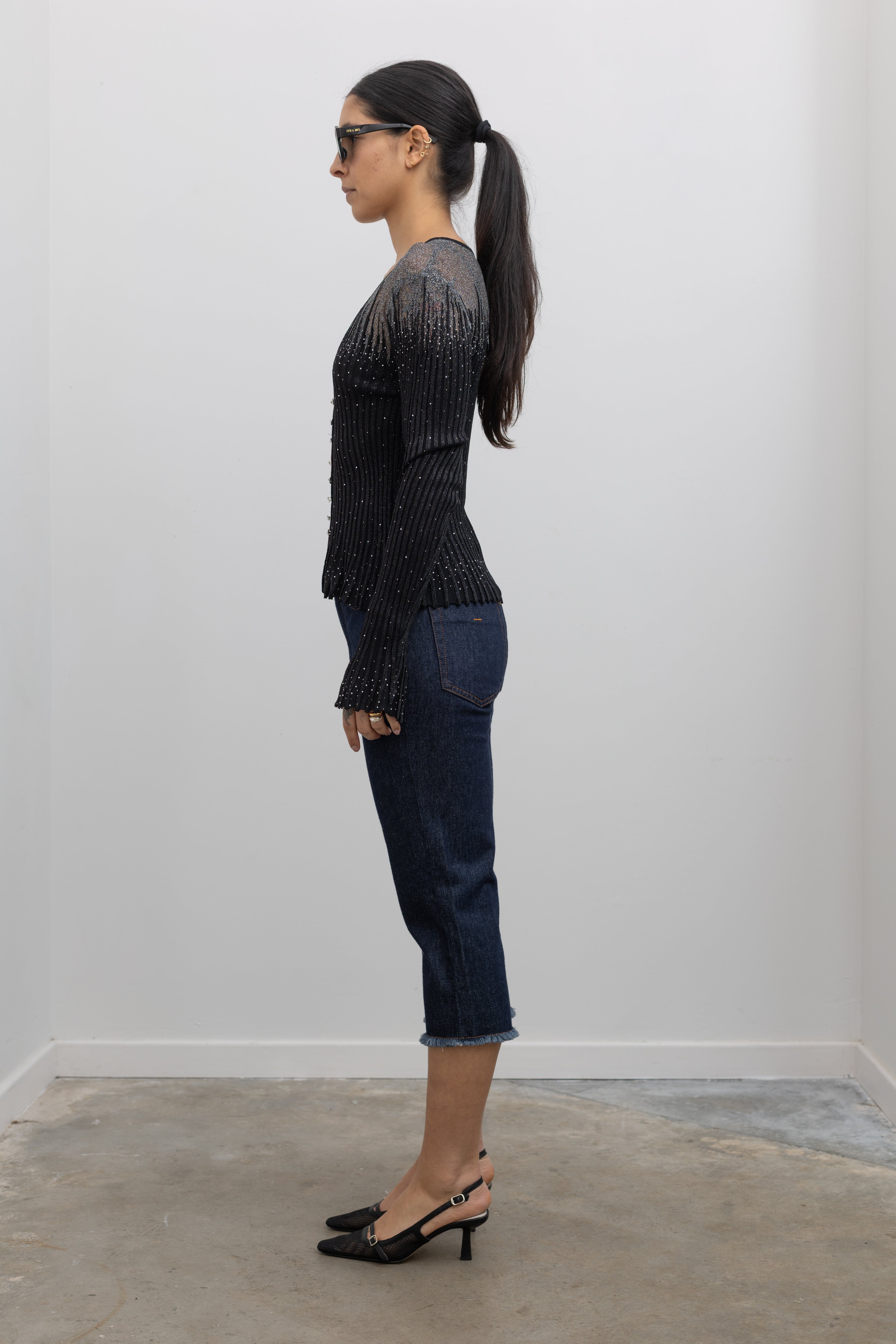 Side view of a woman wearing a Collective Label black top featuring a metallic jeweled motif throughout with long bell sleeves and a faux button down front paired with dark wash denim jeans at an Alas Eius.