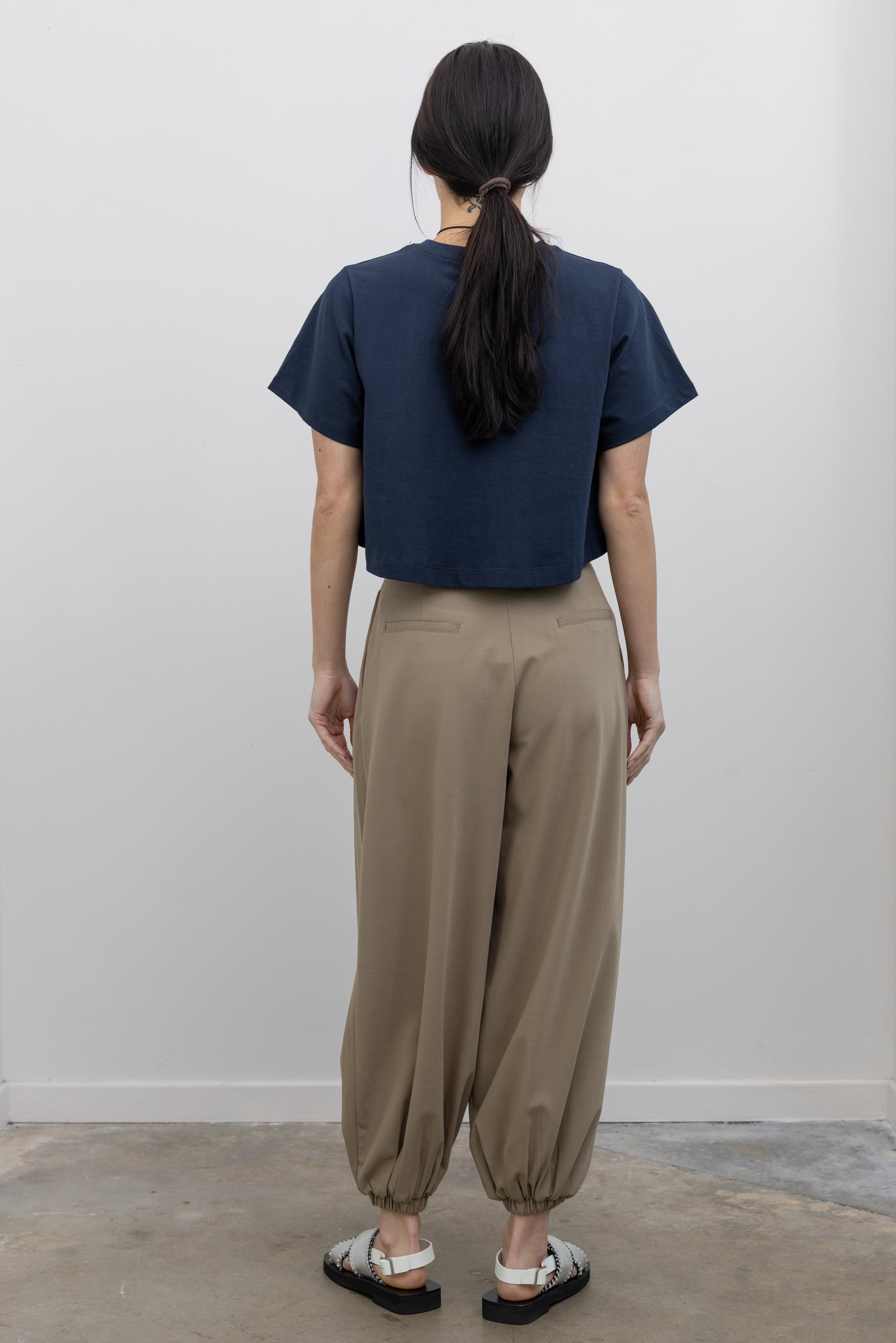 Full length back view of a woman wearing modern minimalist spring fashion in a Collective brand beige Tores pleated front harem pant featuring a cinched elastic ankle creating a balloon silhouette paired with an Alas Eius label navy blue t-shirt and metallic silver sandals at an Alas Eius store.