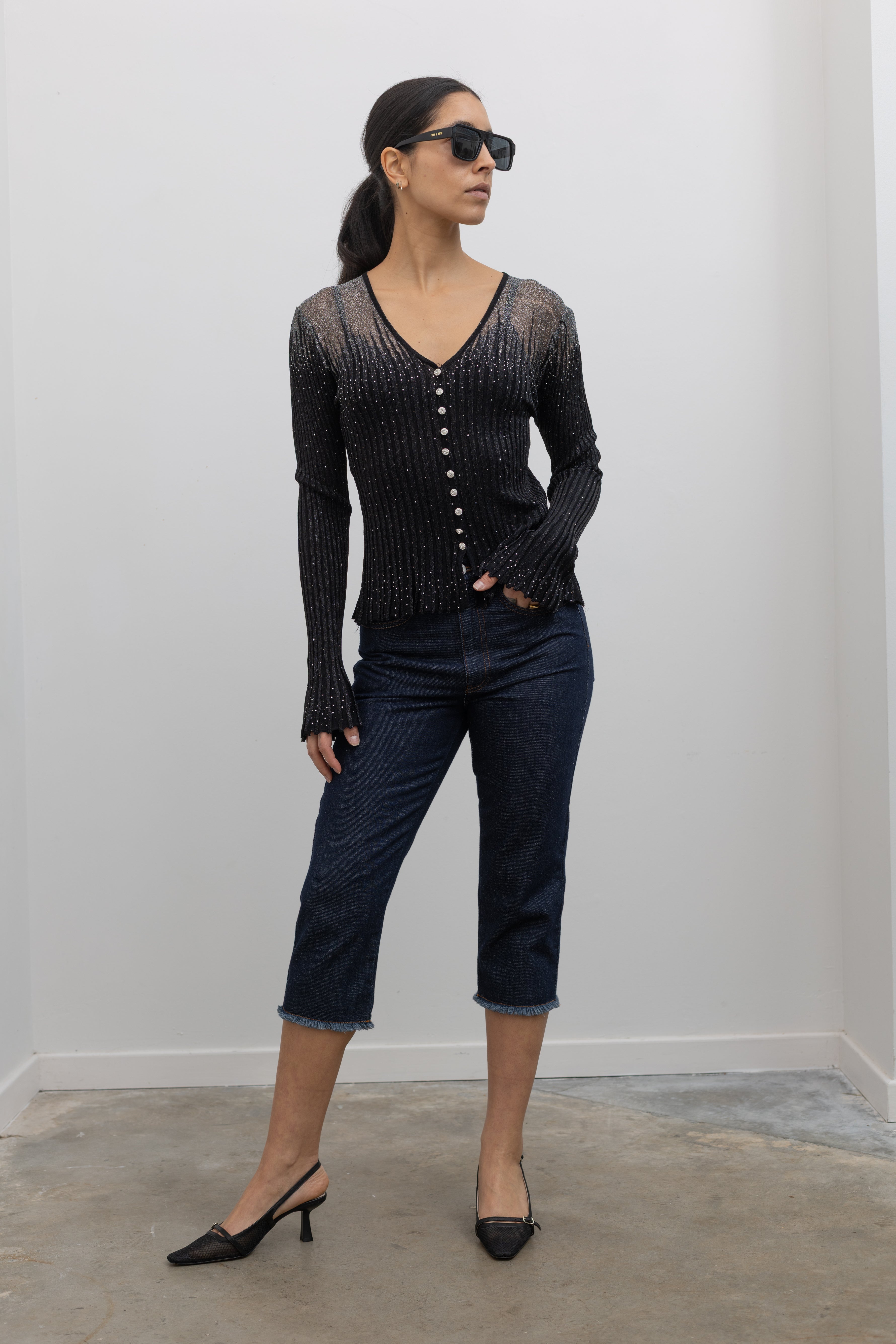 Woman wearing a Collective Label black top featuring a metallic jeweled motif throughout with long bell sleeves and a faux button down front paired with dark wash denim jeans at an Alas Eius.