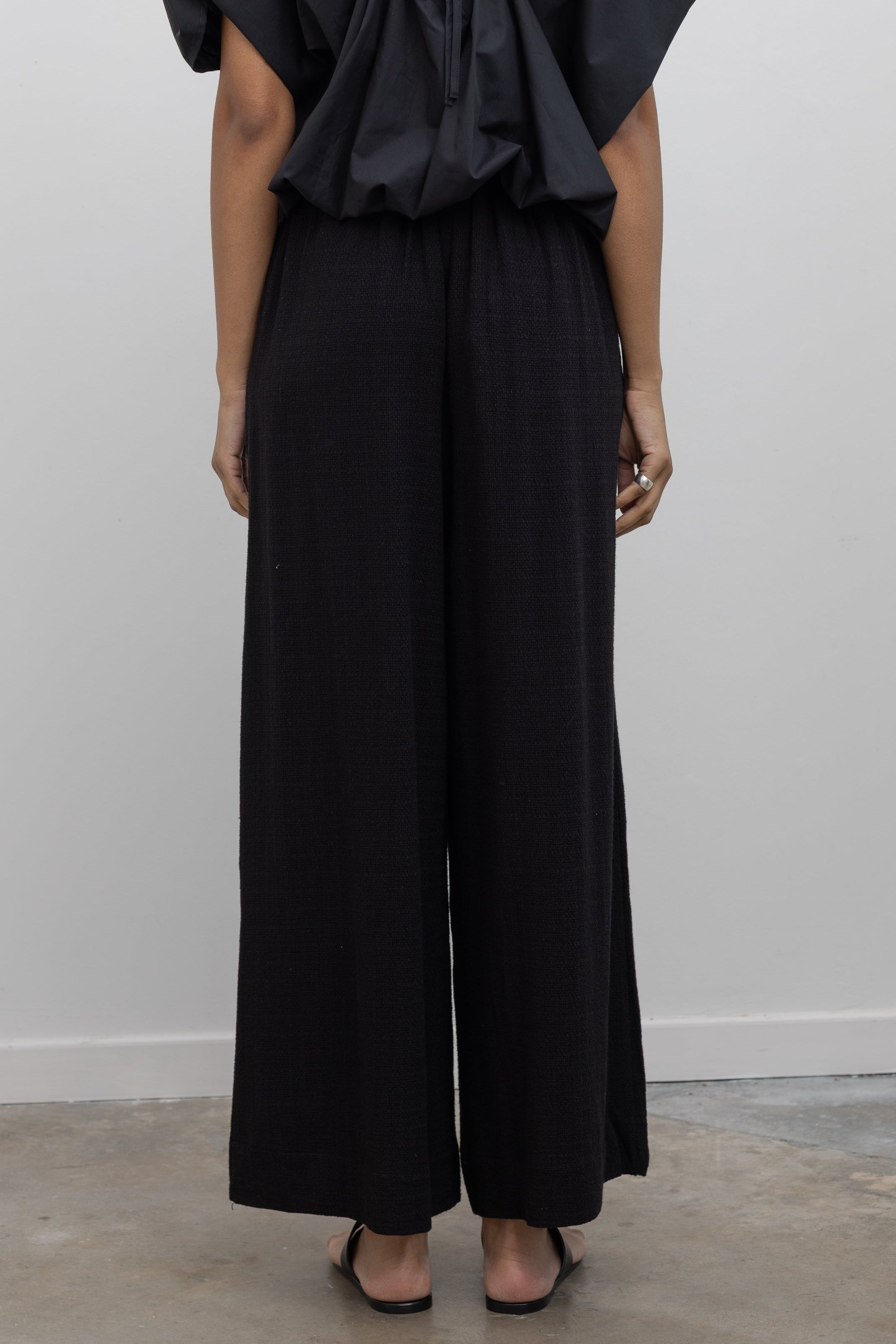 Full length back view of a woman posing wearing casual yet chic spring fashion in the Collective brand black linen wide leg pull on pants with pockets paired with a black balloon hem top and ATP black leather cutout sandals at an Alas Eius store. 