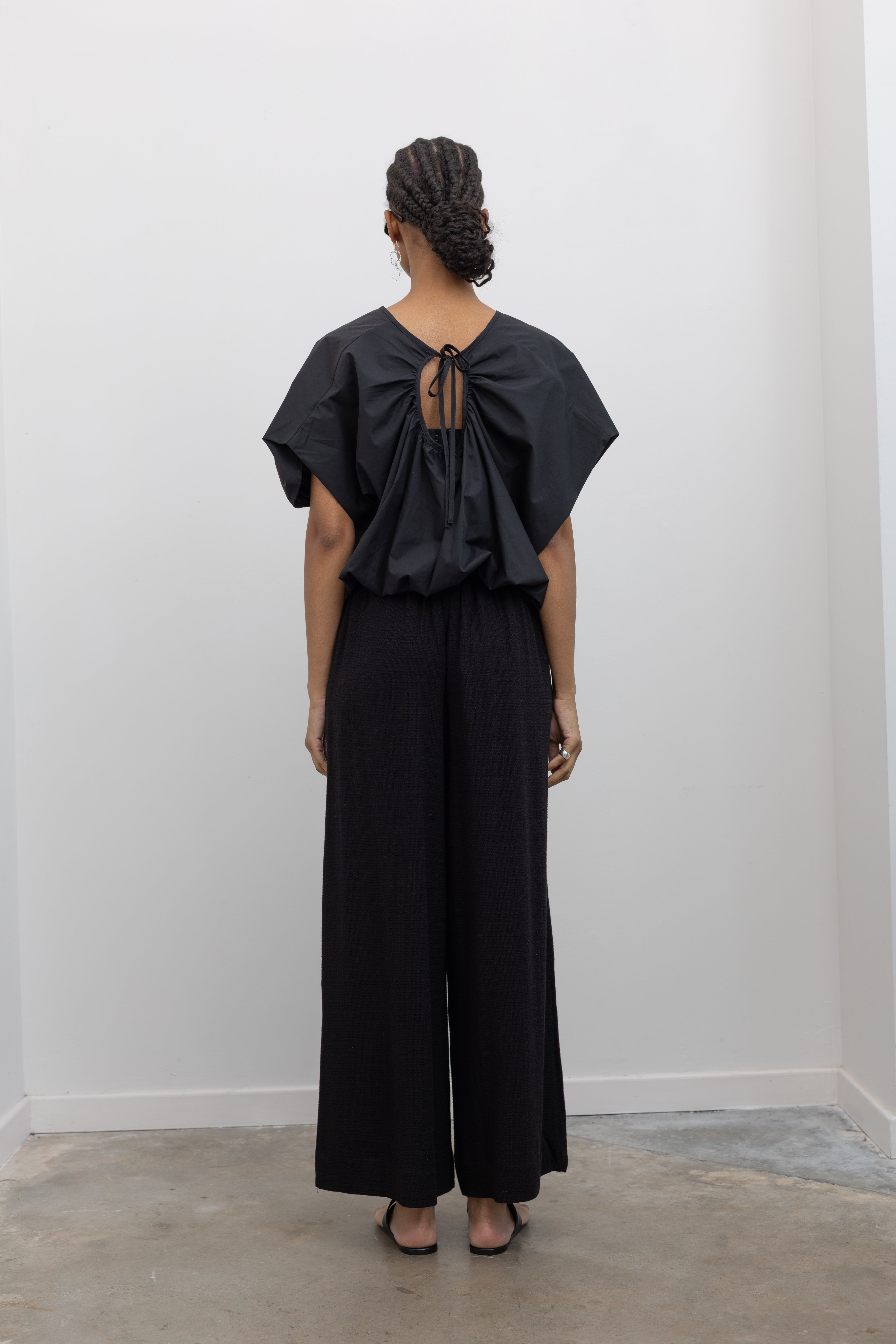 Full length back view of a woman posing wearing casual yet chic spring fashion in the Collective brand black linen wide leg pull on pants with pockets paired with a black balloon hem top and ATP black leather cutout sandals at an Alas Eius store. 