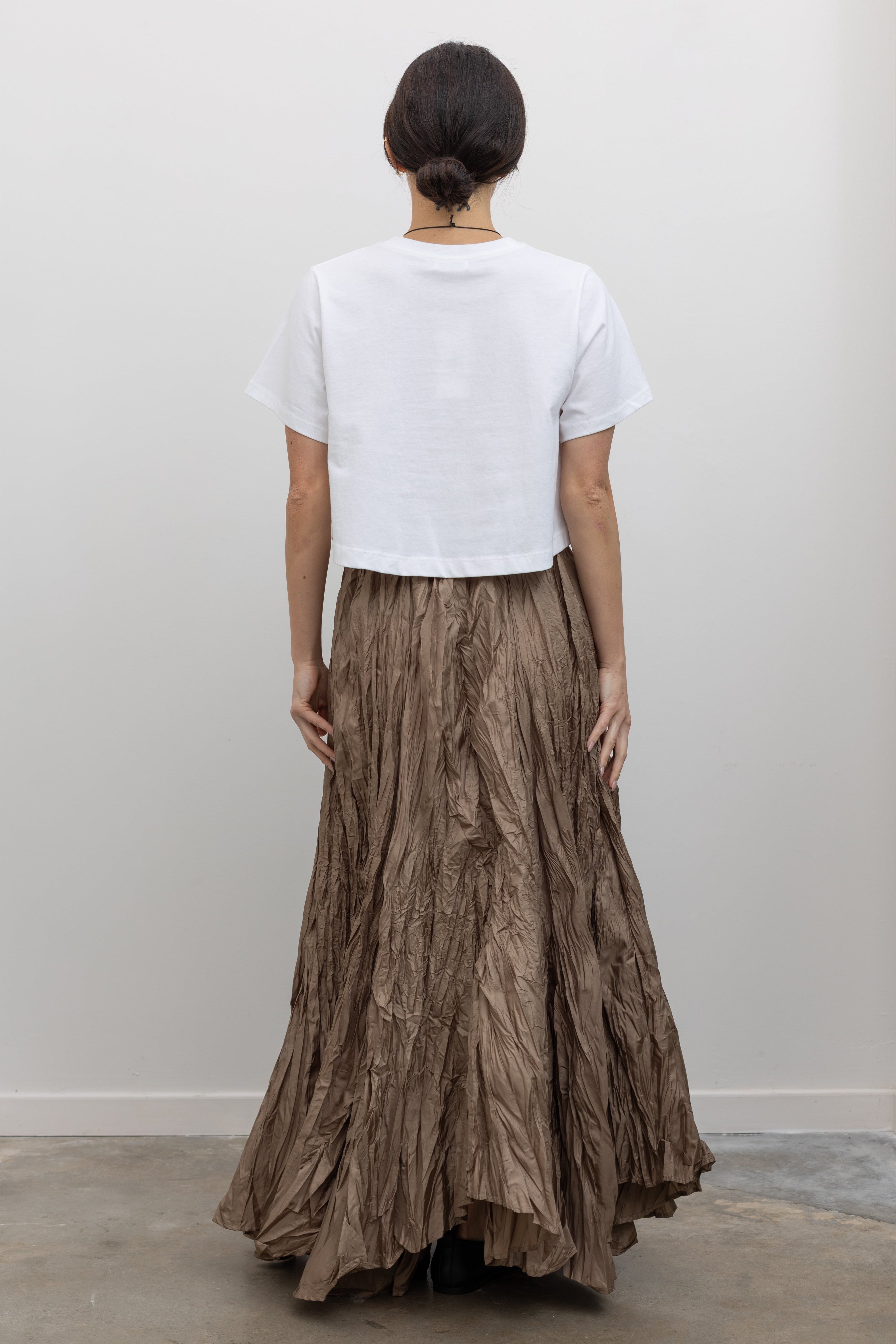 Full length back view of a woman in motion wearing elevated spring fashion in an Alas Eius label cropped white t-shirt paired with a Collective Khaki Crinkle Textured Maxi Skirt at Alas Eius. 