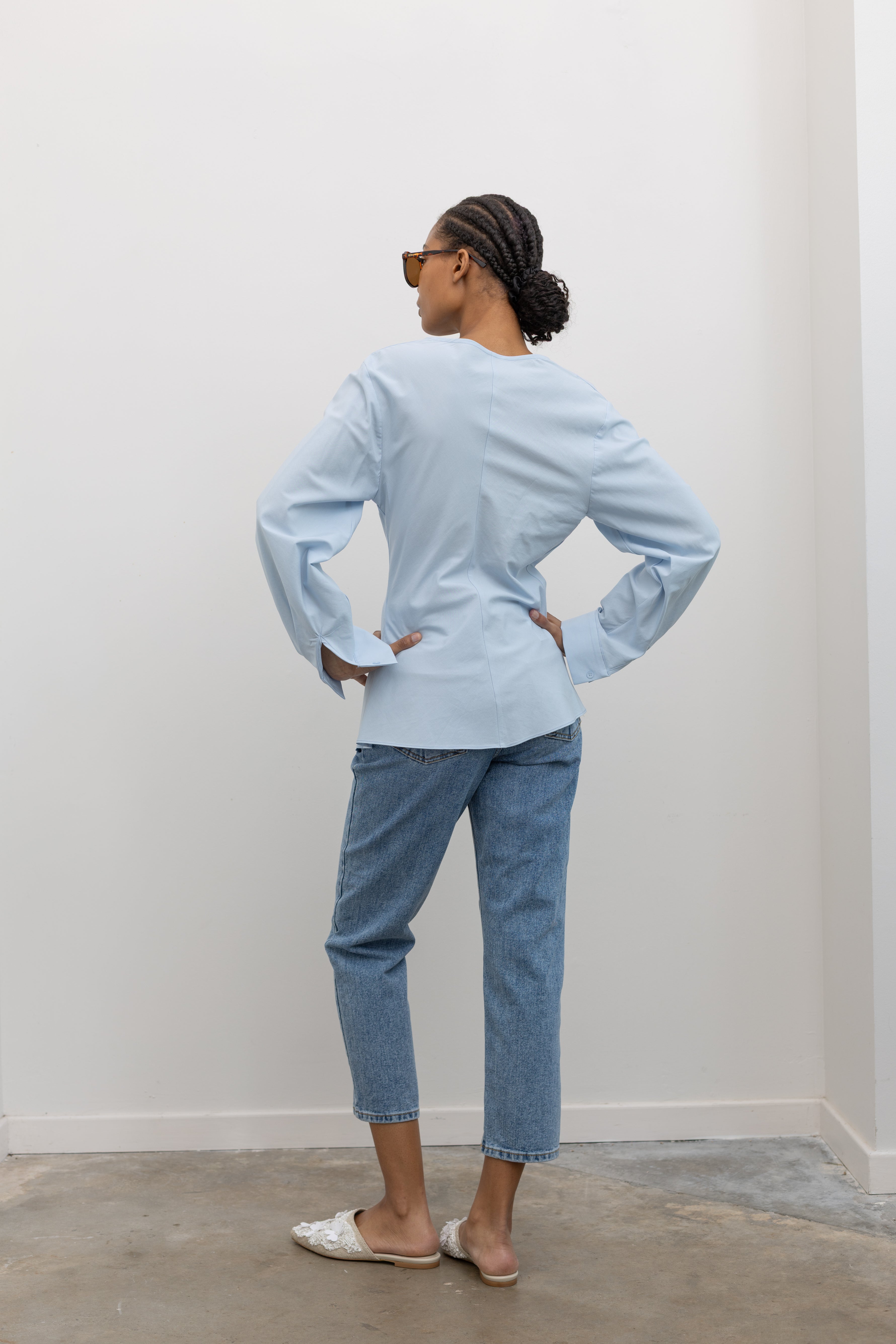 Full length back view of a woman posing wearing chic and architectural spring fashion in a Collective label sky blue cotton long sleeve top featuring a fitted natural waistline with a side draped motif creating an asymmetrical hem and cowl neckline paired with light wash denim jeans at Alas Eius.