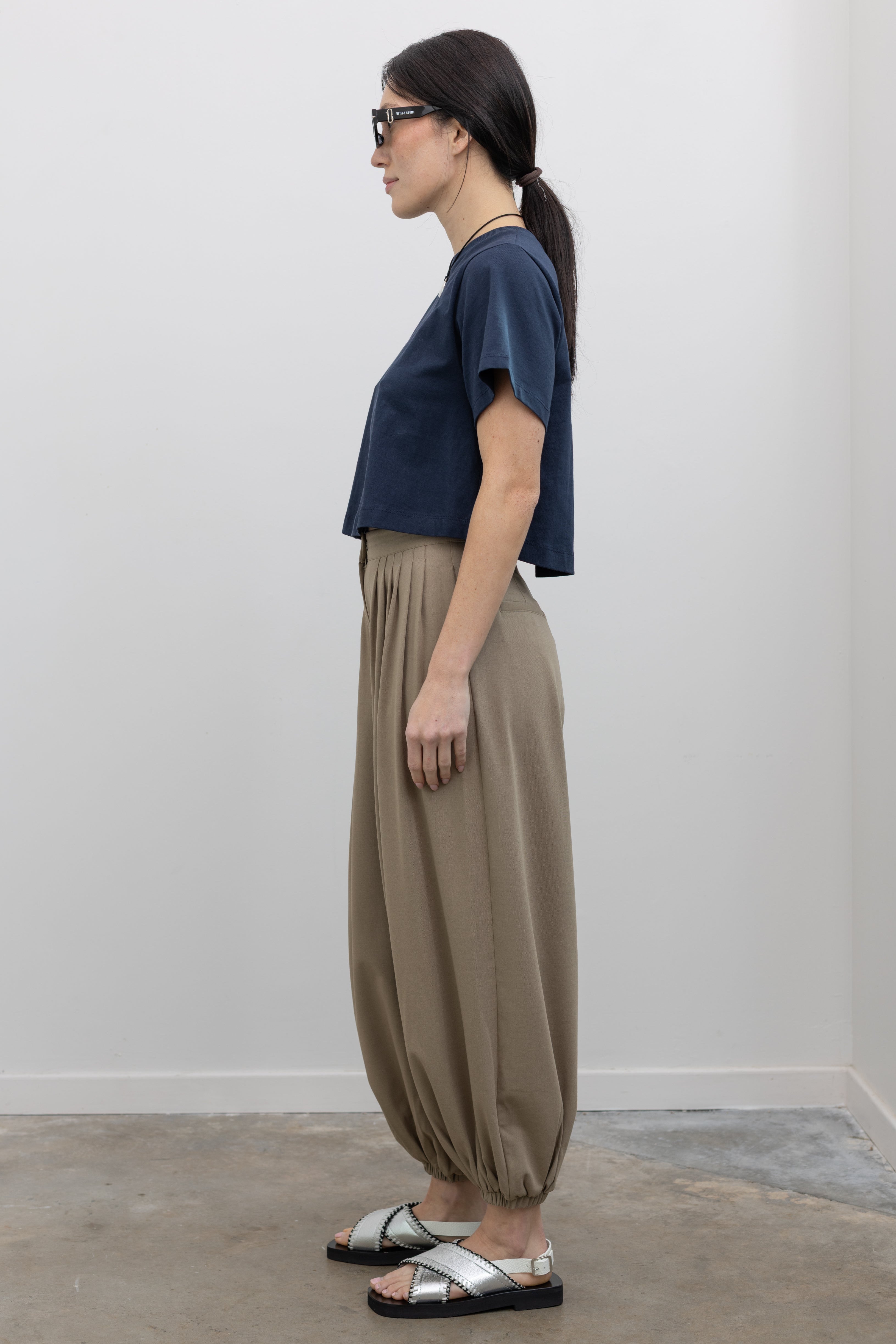 Full length side view of a woman wearing the perfect elevated weekend outfit in a Collective brand beige Tores pleated front harem pant featuring a cinched elastic ankle creating a balloon silhouette paired with an Alas Eius label navy blue t-shirt and metallic silver sandals at an Alas Eius store.