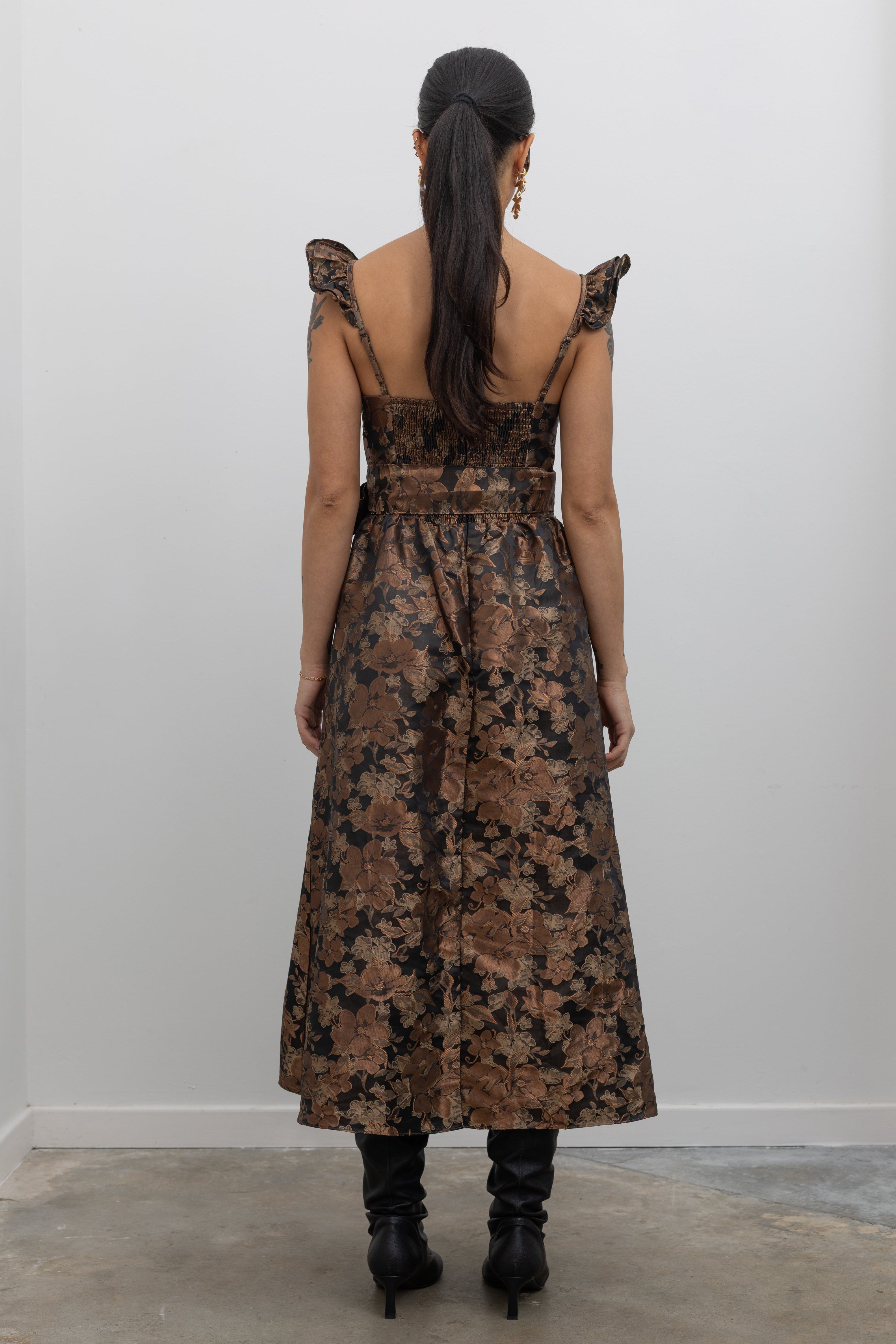 Full back view of a woman wearing flattering feminine Collective label bronze floral jacquard corset midi dress with slouchy black leather heeled boots at Alas Eius. 