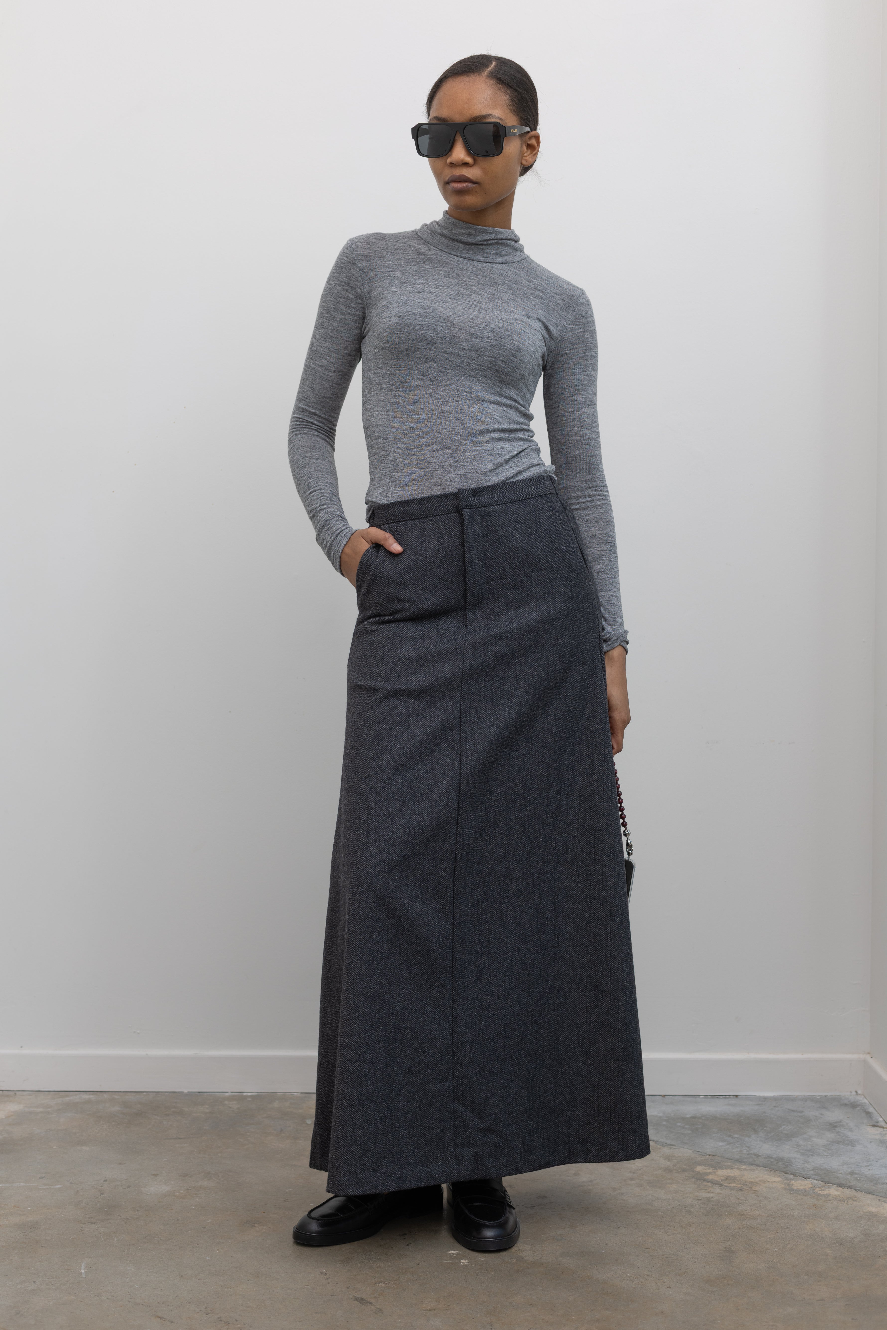 Full length front view of a woman wearing a sophisticated and refined outfit featuring an Collective label charcoal gray herringbone weave A-line maxi skirt paired with a fitted heather gray mock neck top at Alas Eius. 