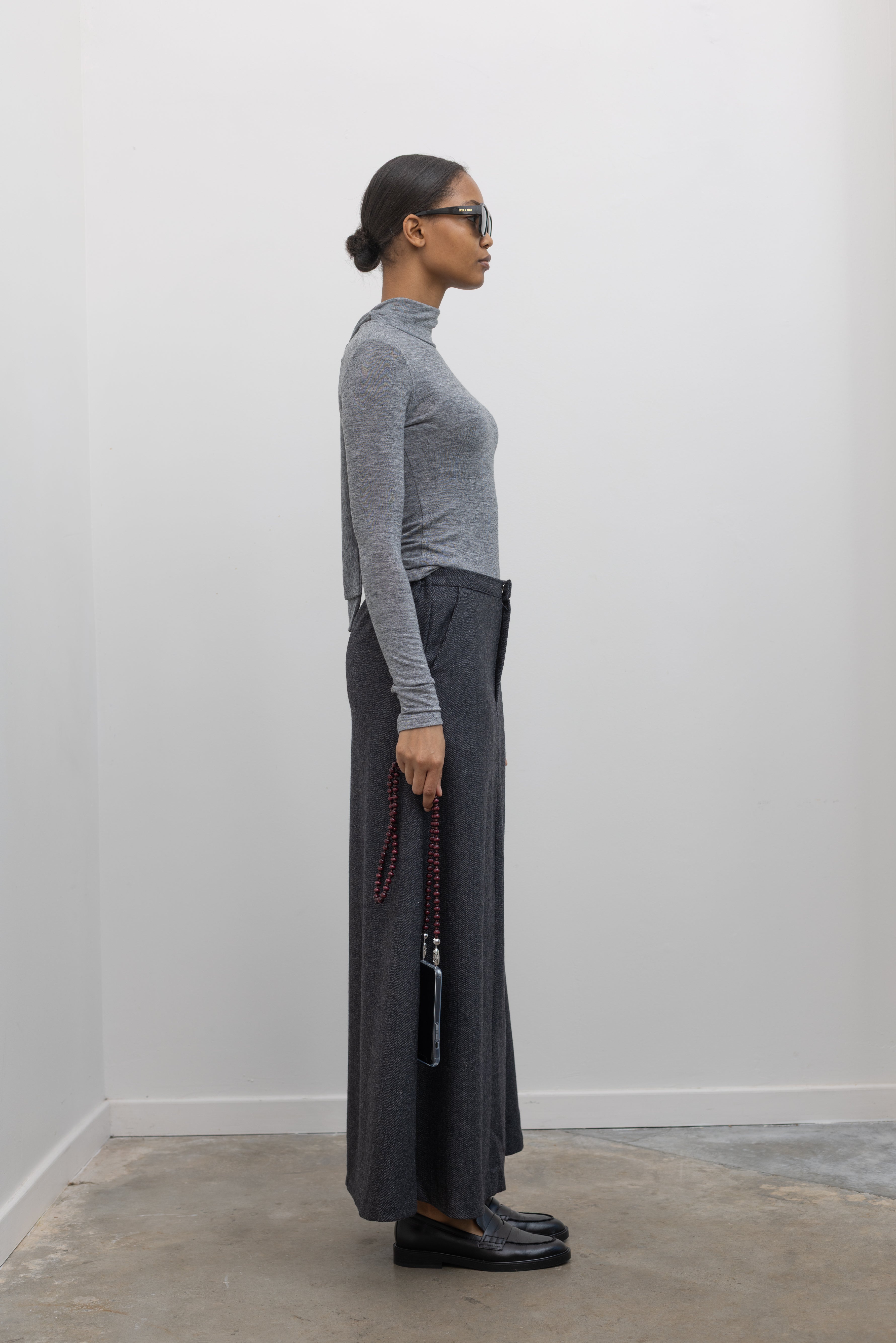 Full length side view of a woman wearing a sophisticated and refined outfit featuring an Collective label charcoal gray herringbone weave A-line maxi skirt paired with a fitted heather gray mock neck top at Alas Eius. 
