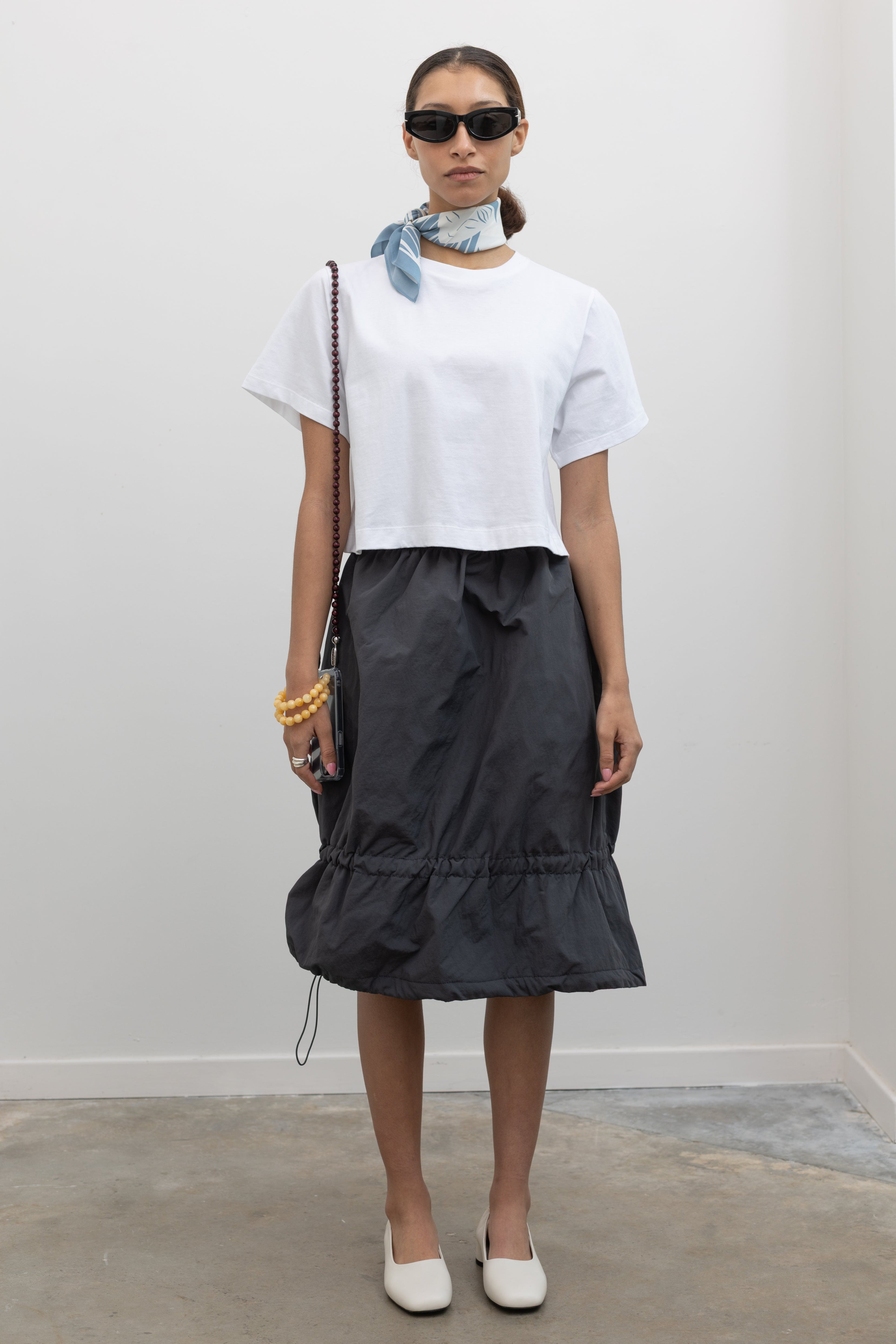 Full front view of a woman standing wearing Spring fashion in a Collective brand charcoal nylon bungee detail midi skirt with an Alas Eius label cropped white t-shirt at Alas Eius. 