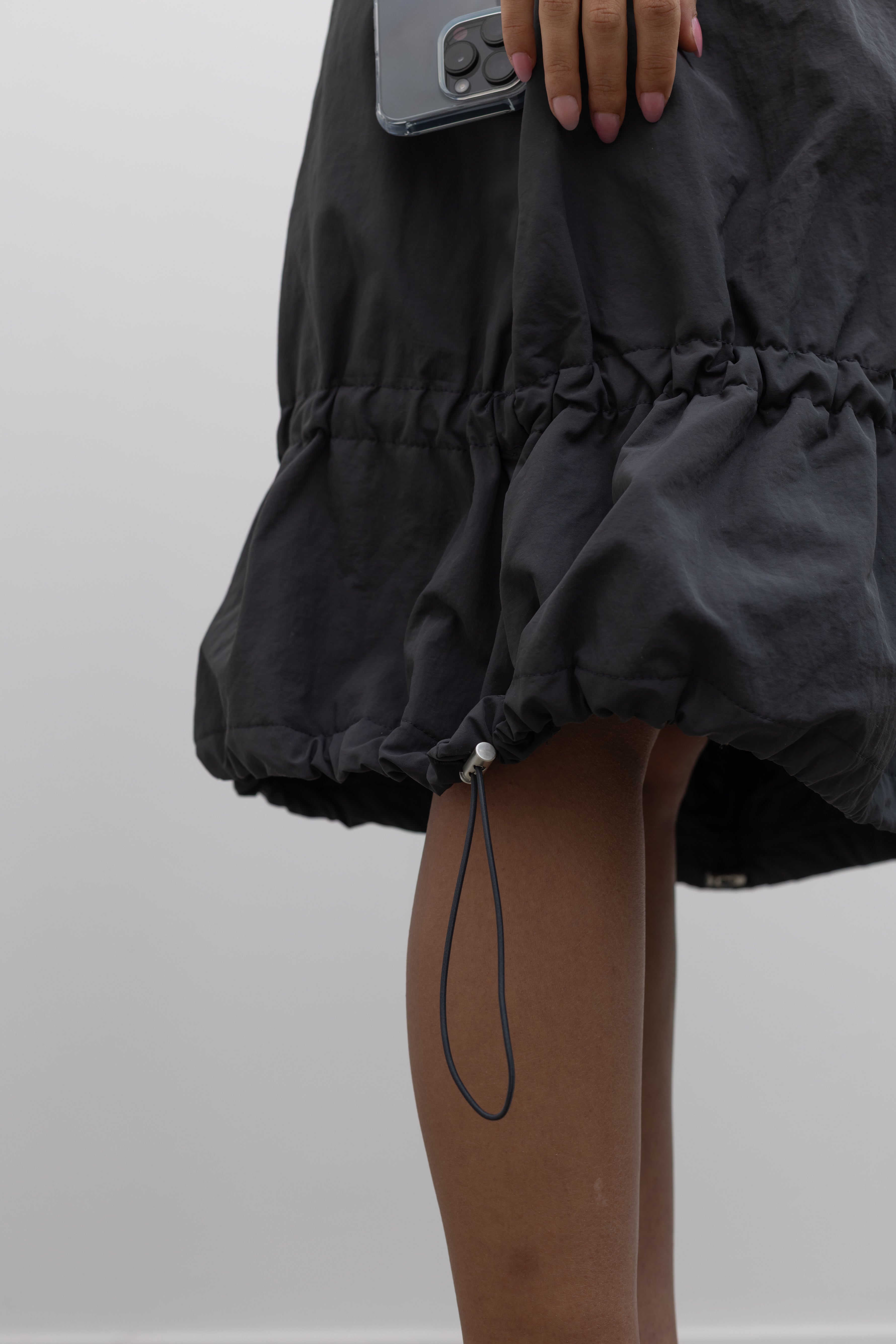 Close-up product view of a woman standing wearing Spring fashion in a sporty Collective brand charcoal nylon bungee cord detail midi skirt at Alas Eius. 