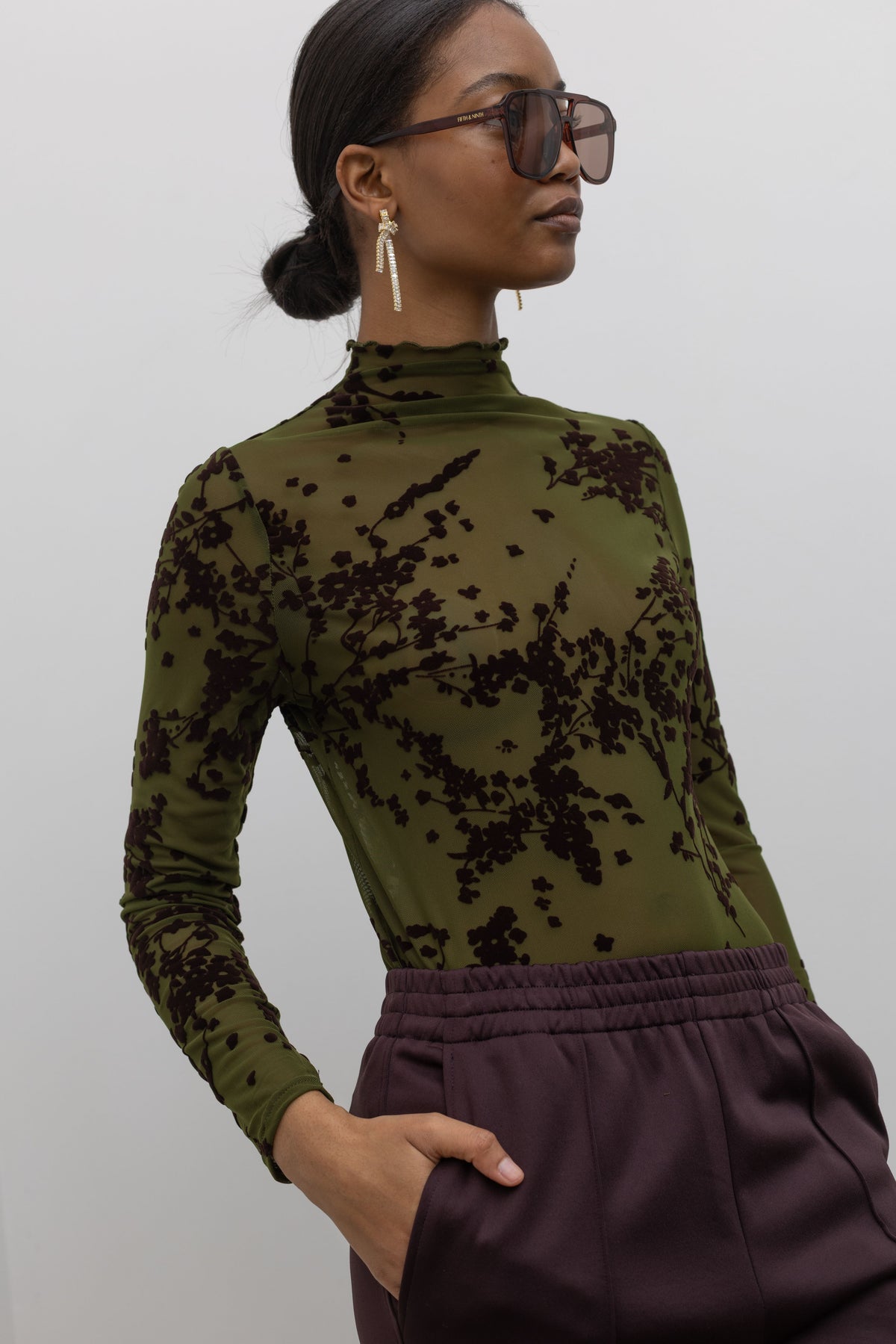 Woman posing wearing a Collective label olive green mesh high neck lettuce hem top featuring a raised floral printed throughout at an Alas Eius store. 