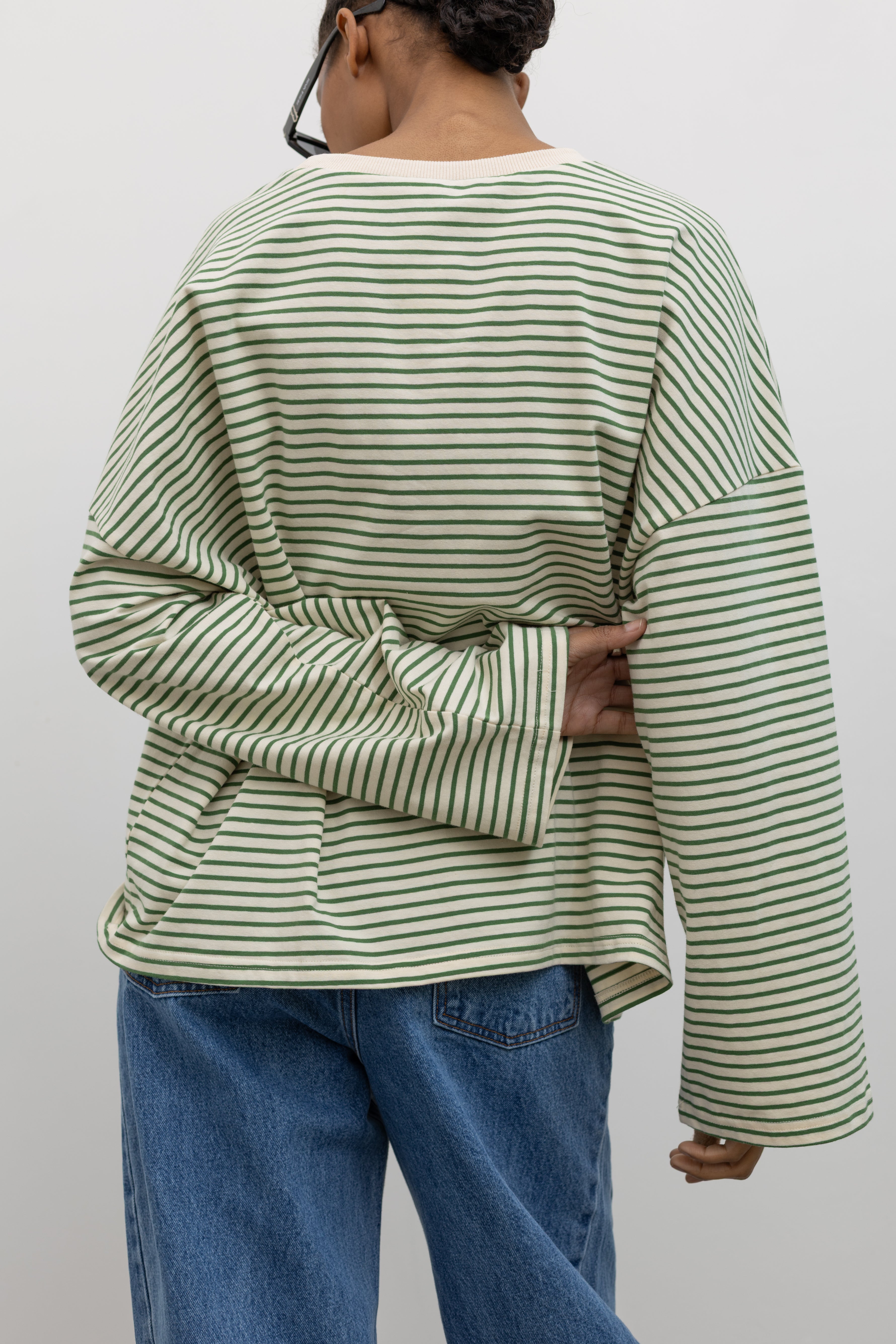 Close-up back view of a woman standing wearing Spring casual fashion in a Collective green and natural stripe dropped shoulder long sleeve cotton  oversized top paired with light wash denim at Alas Eius.