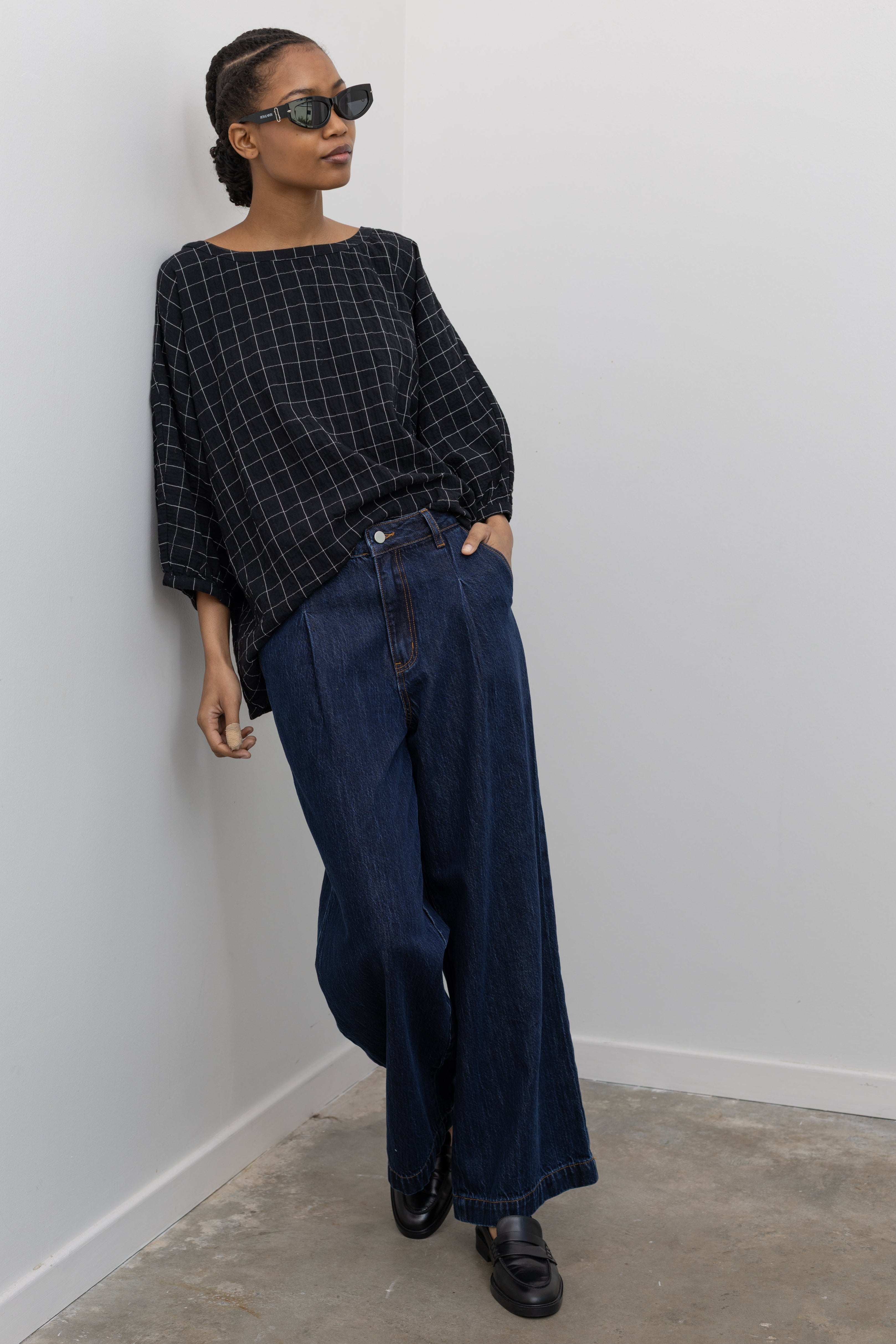 Woman standing leaning against a wall wearing a classic wardrobe staple in a pair of Collective indigo denim wide leg pleated jeans with a black top and black leather loafers at Alas Eius.