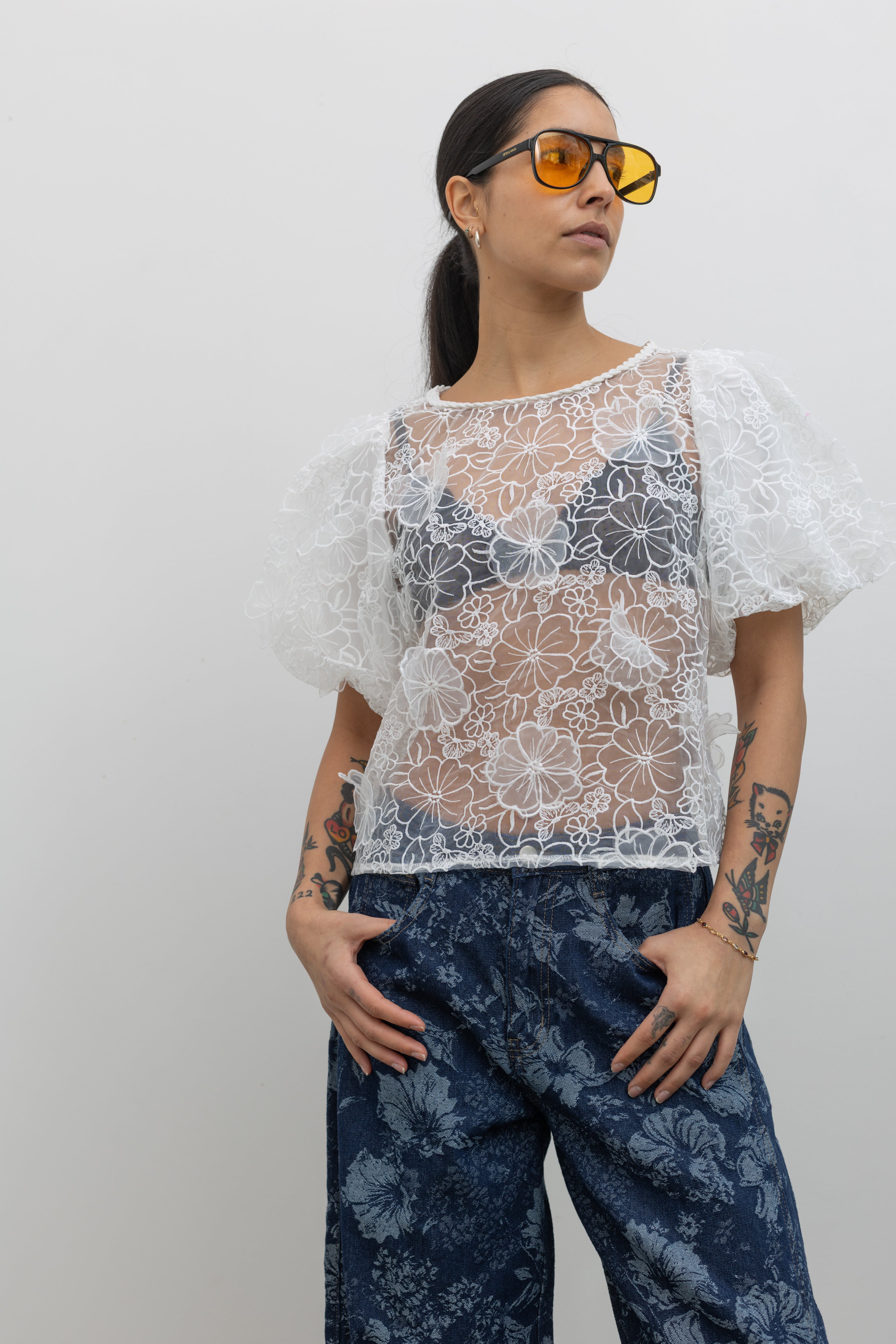 Woman posing wearing Spring fashion in a Collective Ivory Floral Embroidered Billow Sleeve Organza Blouse paired with floral printed dark wash denim jeans at an Alas Eius store. 