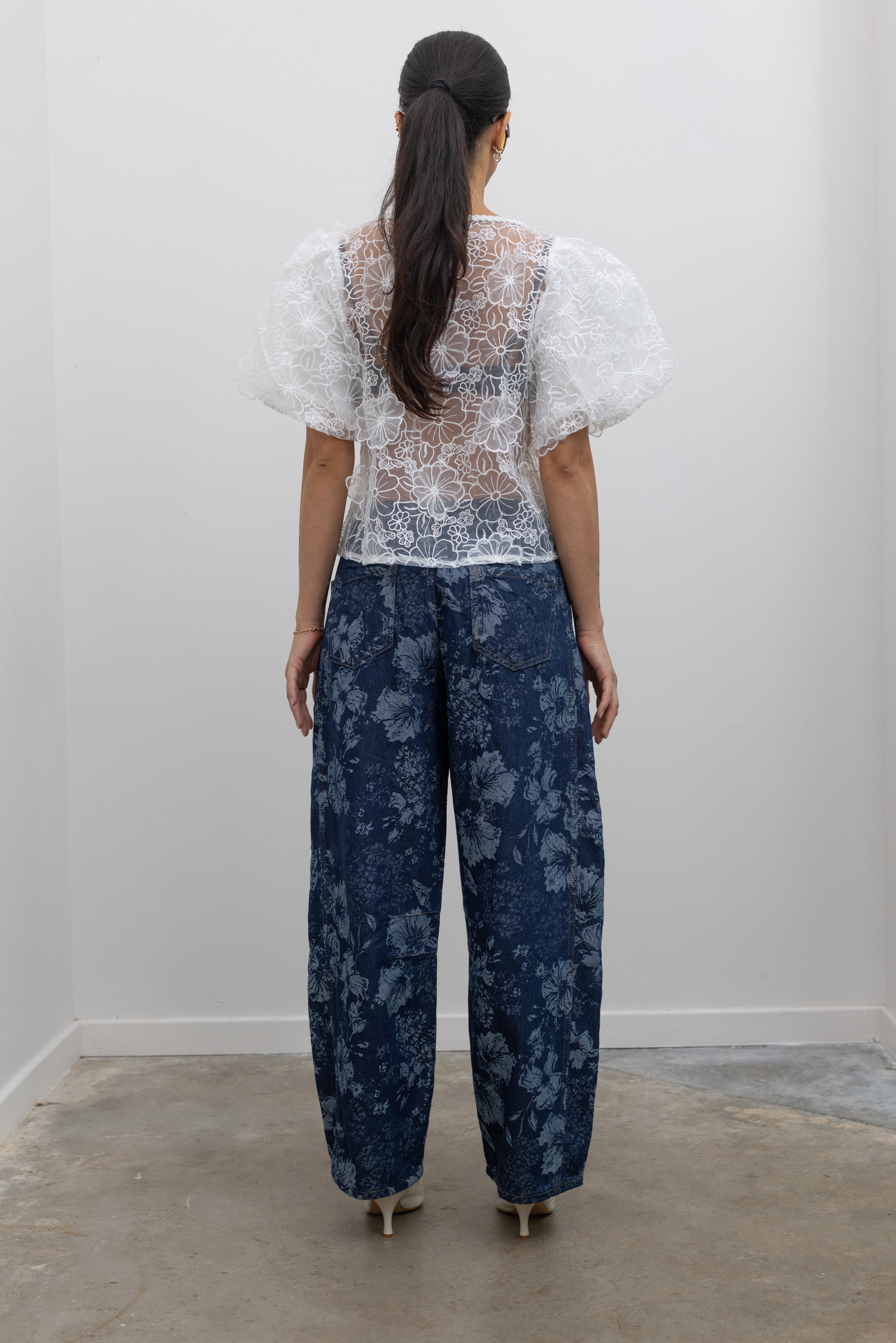 Full length back view of a woman posing wearing Spring fashion in a Collective Ivory Floral Embroidered Billow Sleeve Organza Blouse featuring a semi-sheer front paired with floral printed dark wash denim jeans at an Alas Eius store. 