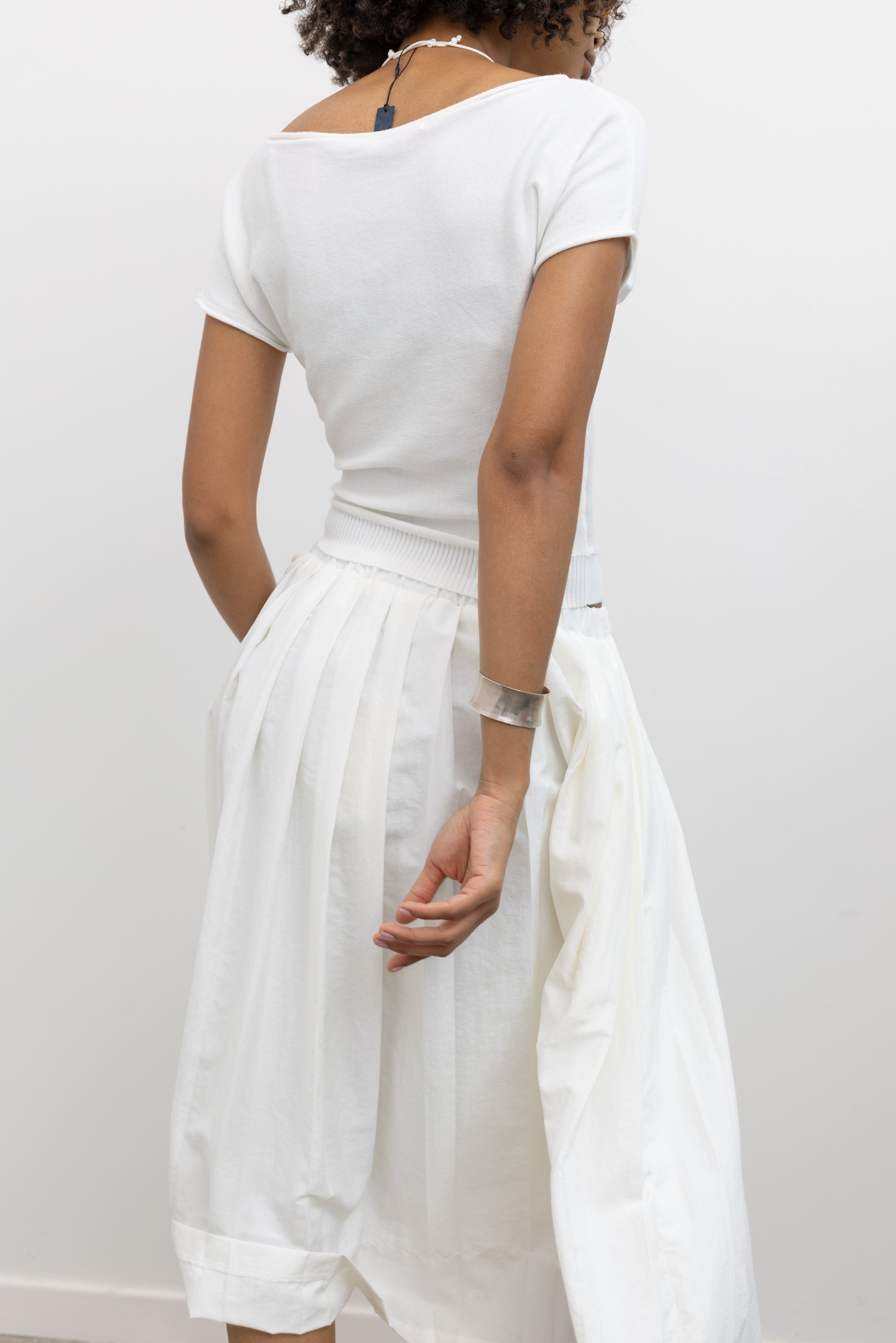 Woman wearing chic summer fashion in a Collective brand ivory knit boat neck cinched tee paired with an Alas Eius label white nylon pleated midi skirt at Alas Eius. 