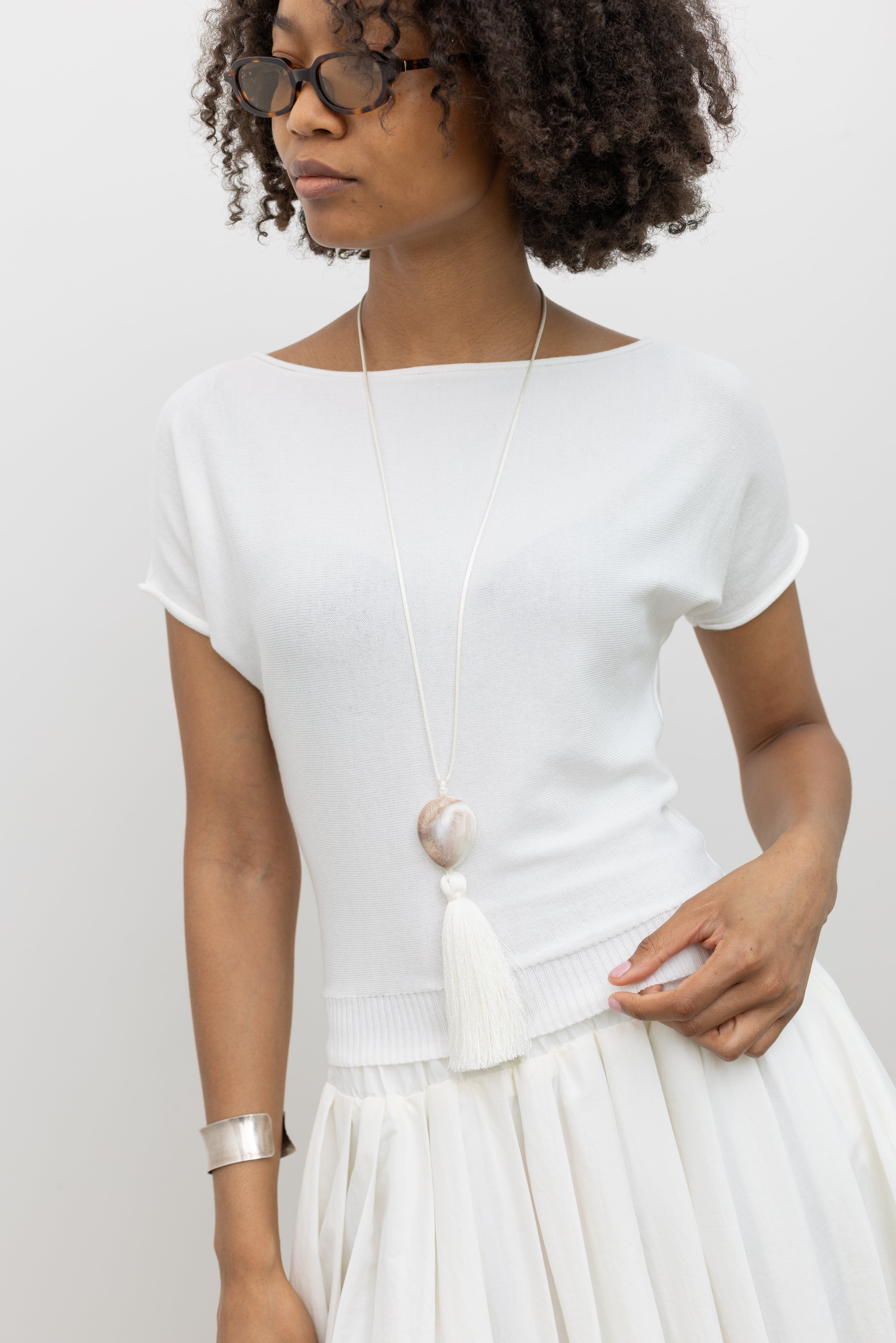 Woman wearing chic summer fashion in a Collective brand ivory knit boat neck cinched tee paired with an Alas Eius label white nylon pleated midi skirt at Alas Eius.   