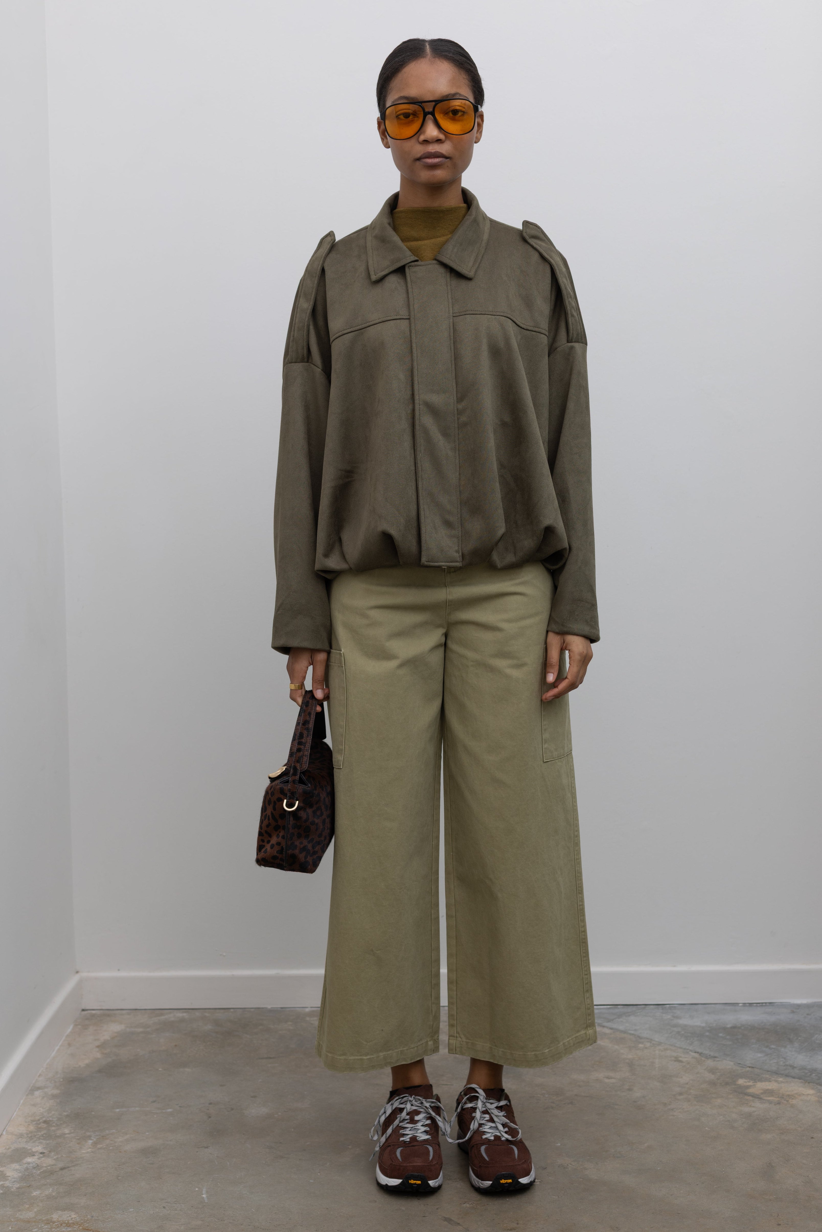 Full length front view of a woman standing wearing a casual weekend outfit in a Collective label khaki green cotton straight leg cropped length cargo style pant paired with a zip jacket and brown sneakers at Alas Eius. 