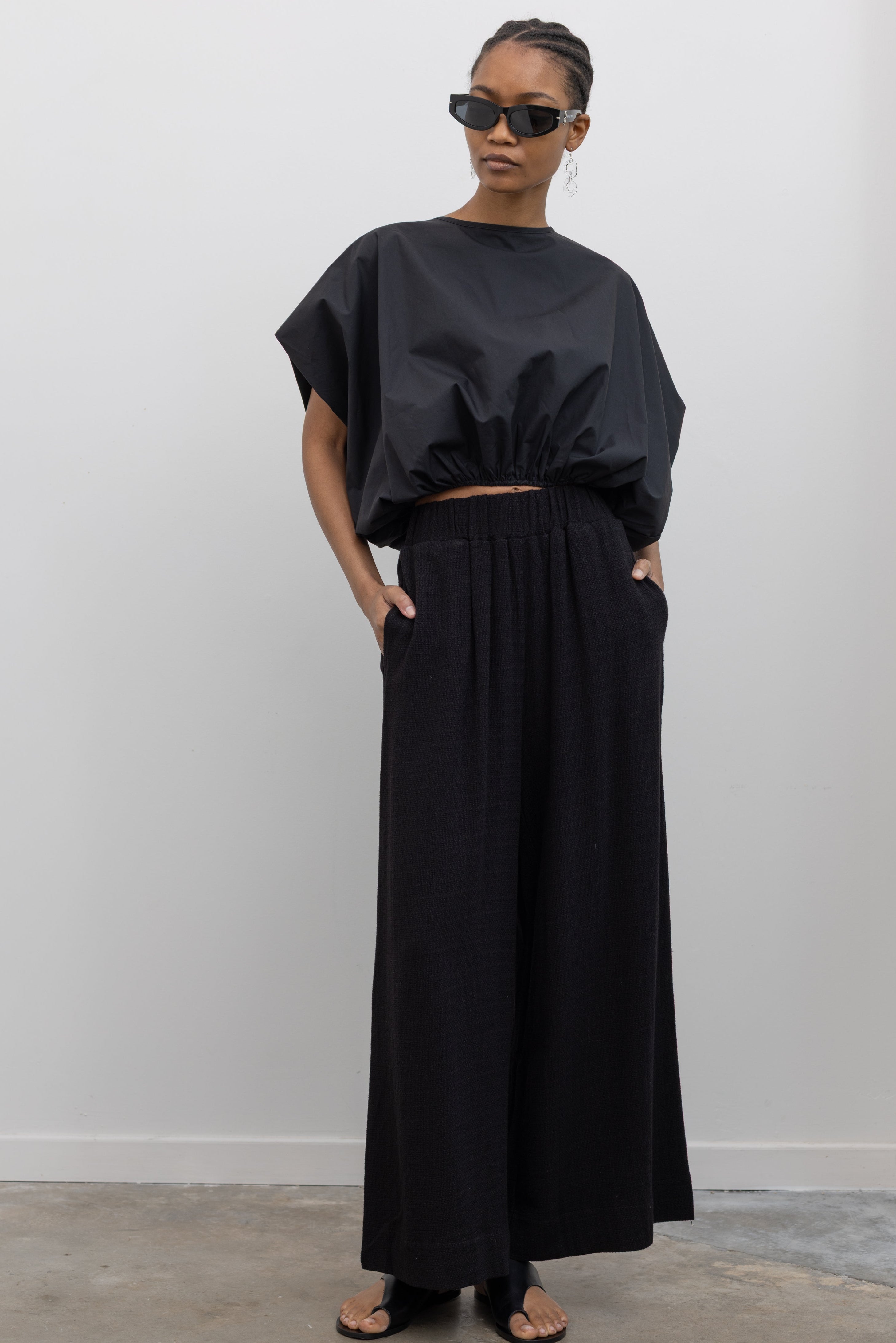 Full length front view of a woman posing wearing contemporary spring fashion in the Collective brand black linen wide leg pull on pants with pockets paired with a black balloon hem top and ATP black leather cutout sandals at an Alas Eius store. 