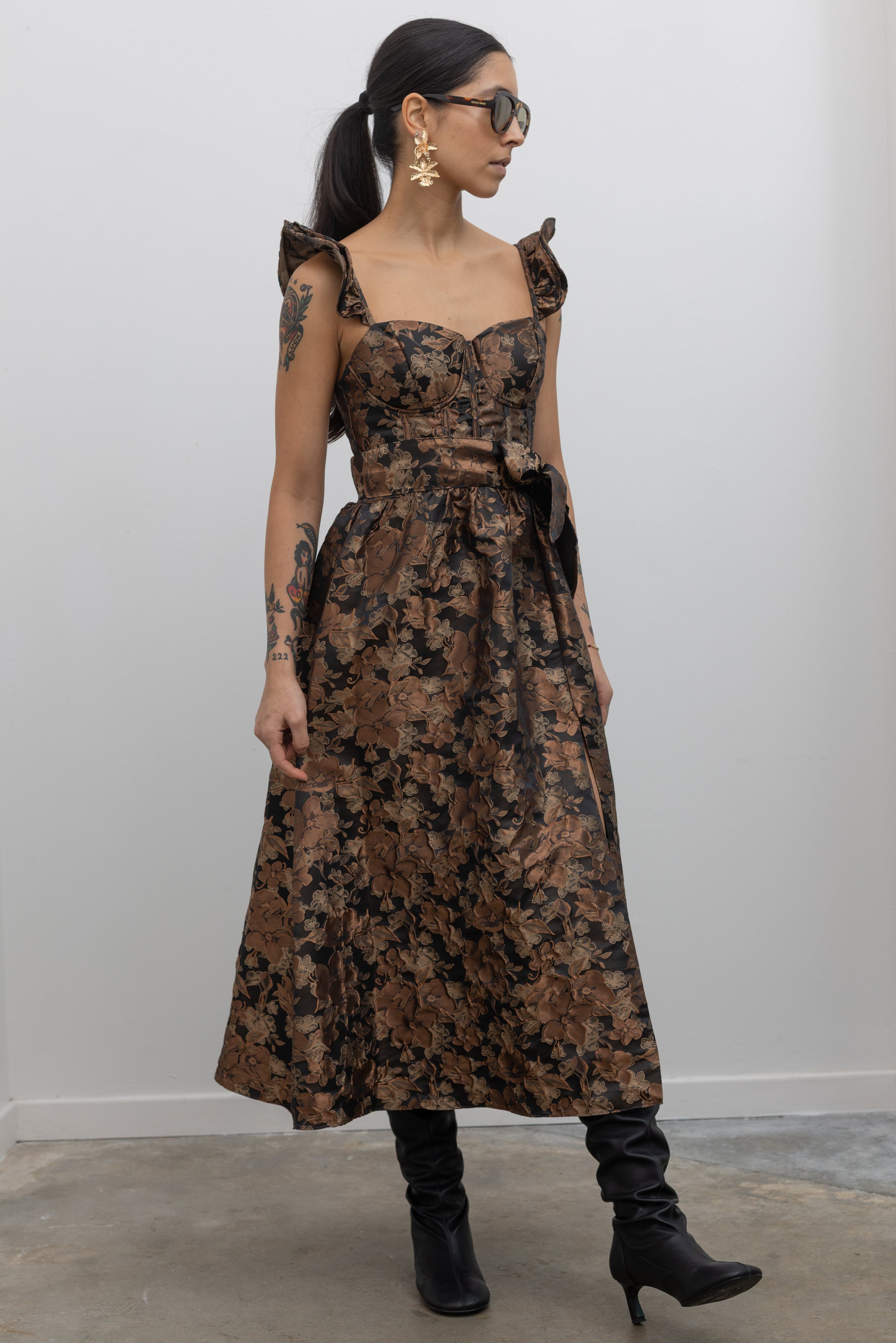 Full front view of a woman in motion wearing a flattering feminine Collective label bronze floral jacquard corset midi dress with slouchy black leather heeled boots at Alas Eius. 