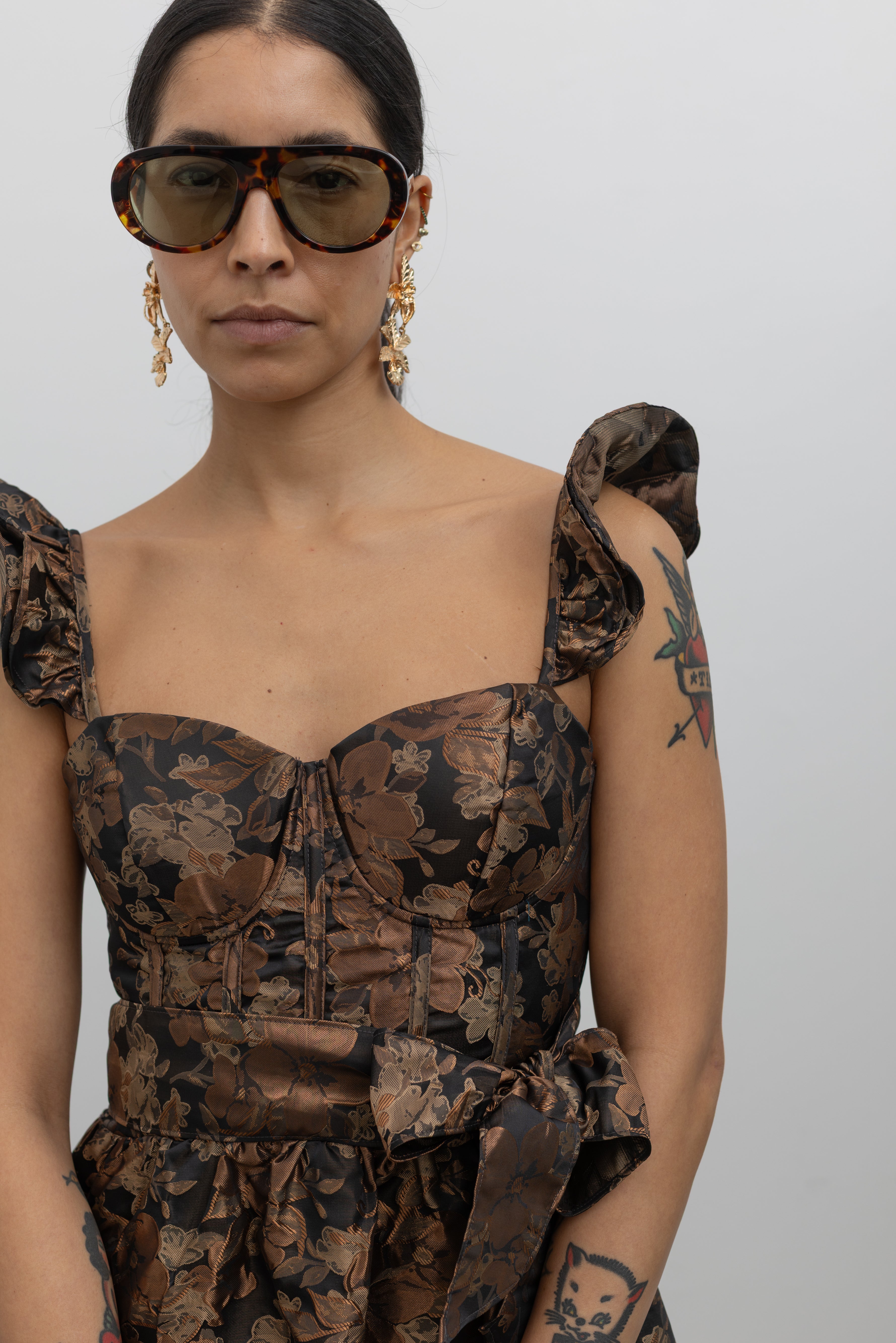 A close-up view of a woman posing wearing an elegant Collective label bronze floral jacquard corset midi dress featuring feminine ruffle straps and a removeable waist bow sash at Alas Eius.