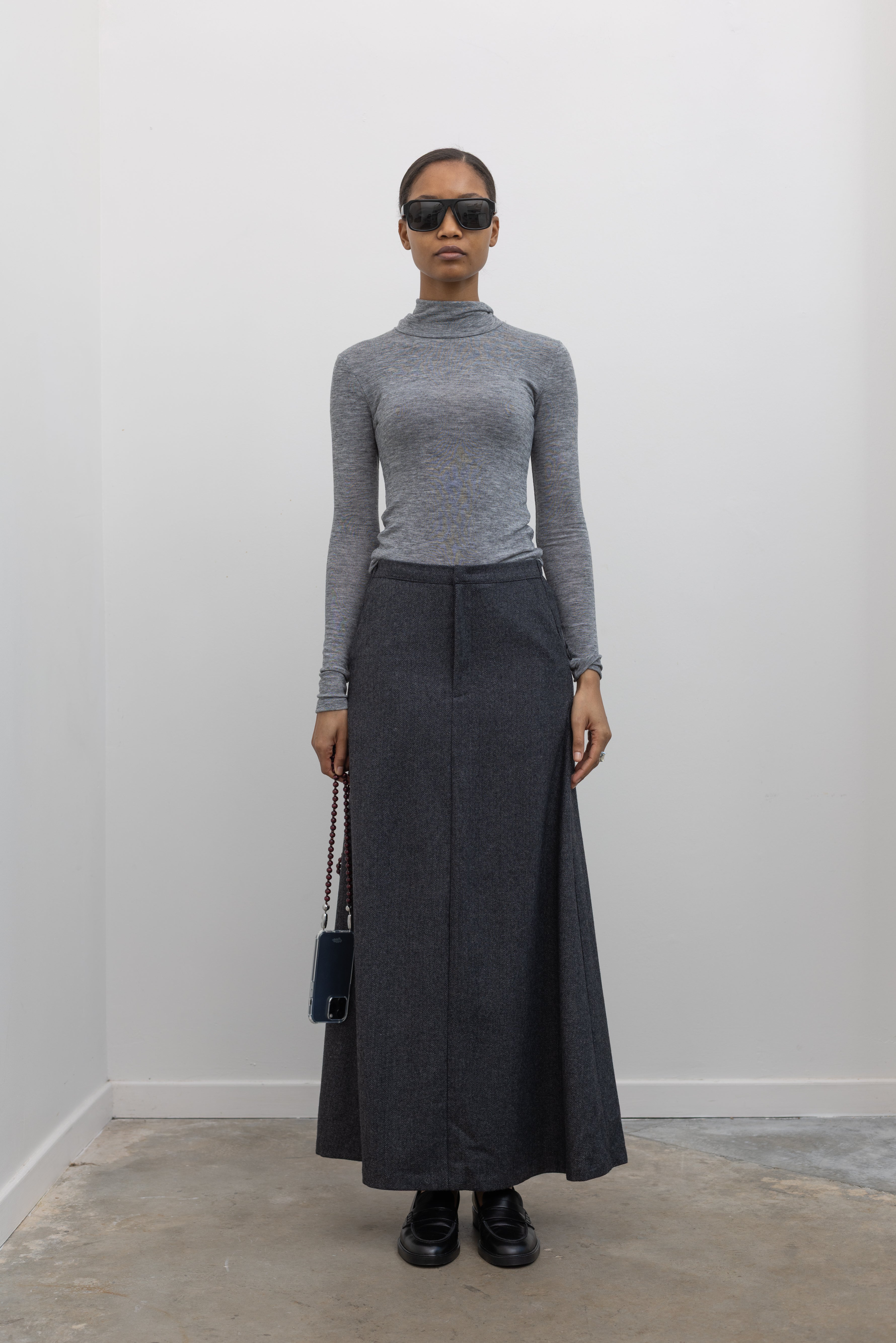 Full length front view of a woman wearing a sophisticated and refined outfit featuring an Collective label charcoal gray herringbone weave A-line maxi skirt paired with a fitted heather gray mock neck top at Alas Eius. 