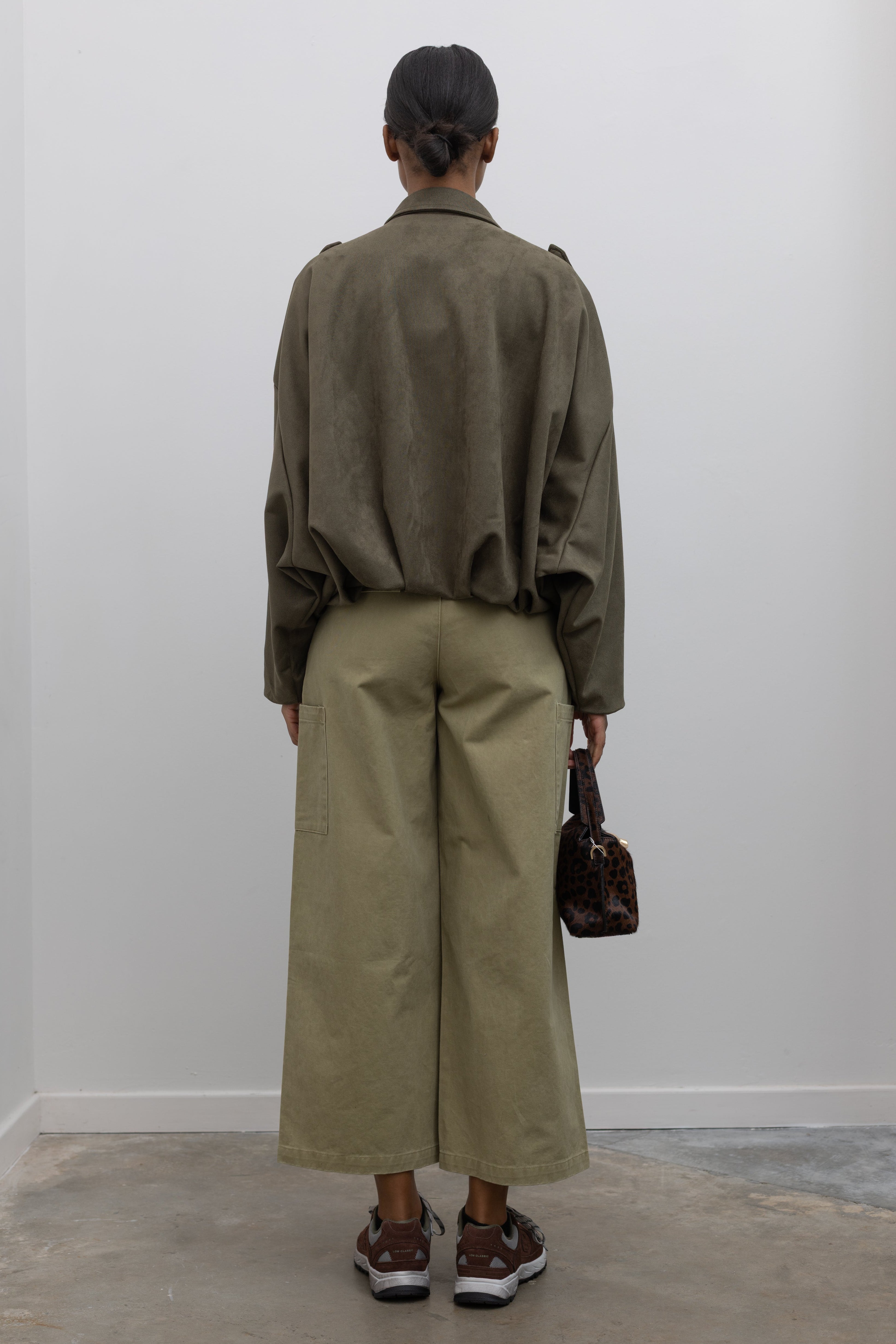 Full length back view of a woman standing wearing a casual weekend outfit in a Collective label khaki green cotton straight leg cropped length cargo style pant paired with a zip jacket and brown sneakers at Alas Eius. 