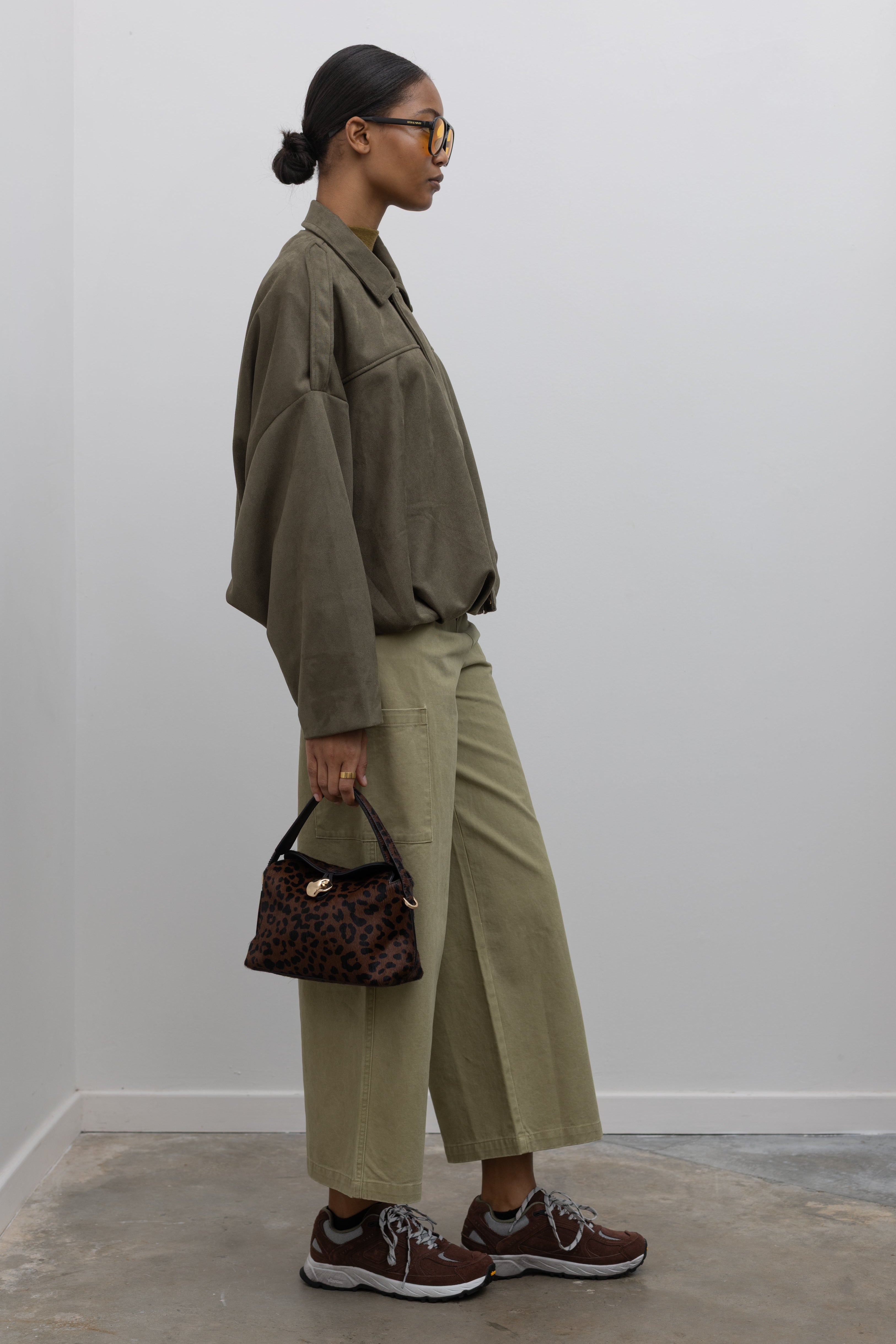 Full length side view of a woman standing wearing a casual weekend outfit in a Collective label khaki green cotton straight leg cropped length cargo style pant paired with a zip jacket and brown sneakers at Alas Eius. 