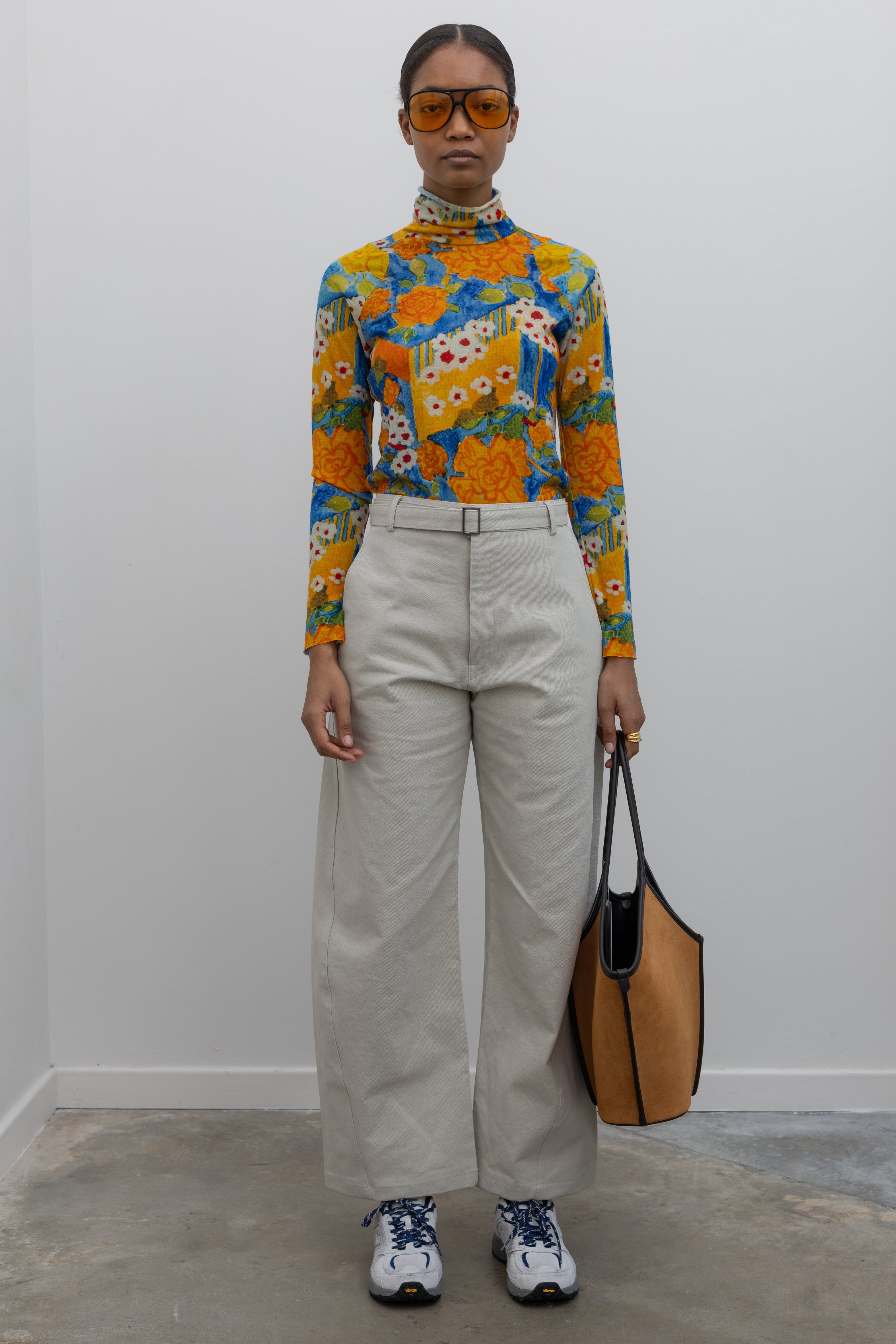 Front view of a woman wearing weekend fashion in a pair of Collective brand light gray curved leg belted chino pants with a vibrant orange and blue floral printed long sleeve top paired with white and blue sneakers at Alas Eius.
