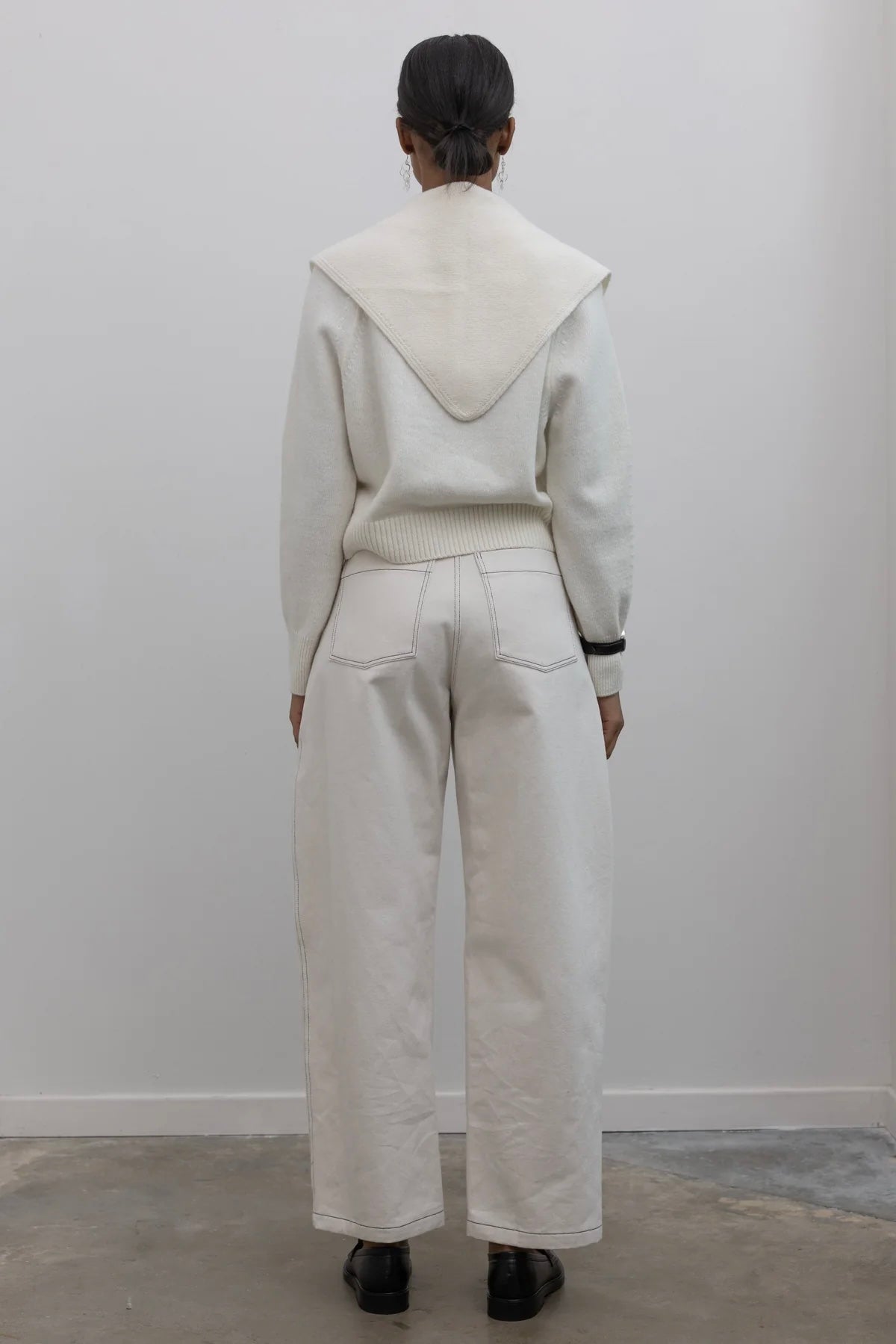 Full length back view of a woman standing wearing an everyday capsule wardrobe outfit in a white long sleeve top with Collective label Spring Cream Curved leg pants featuring Contrasting stitching paired with a black leather belt with silver hardware and black leather loafers at Alas Eius. 