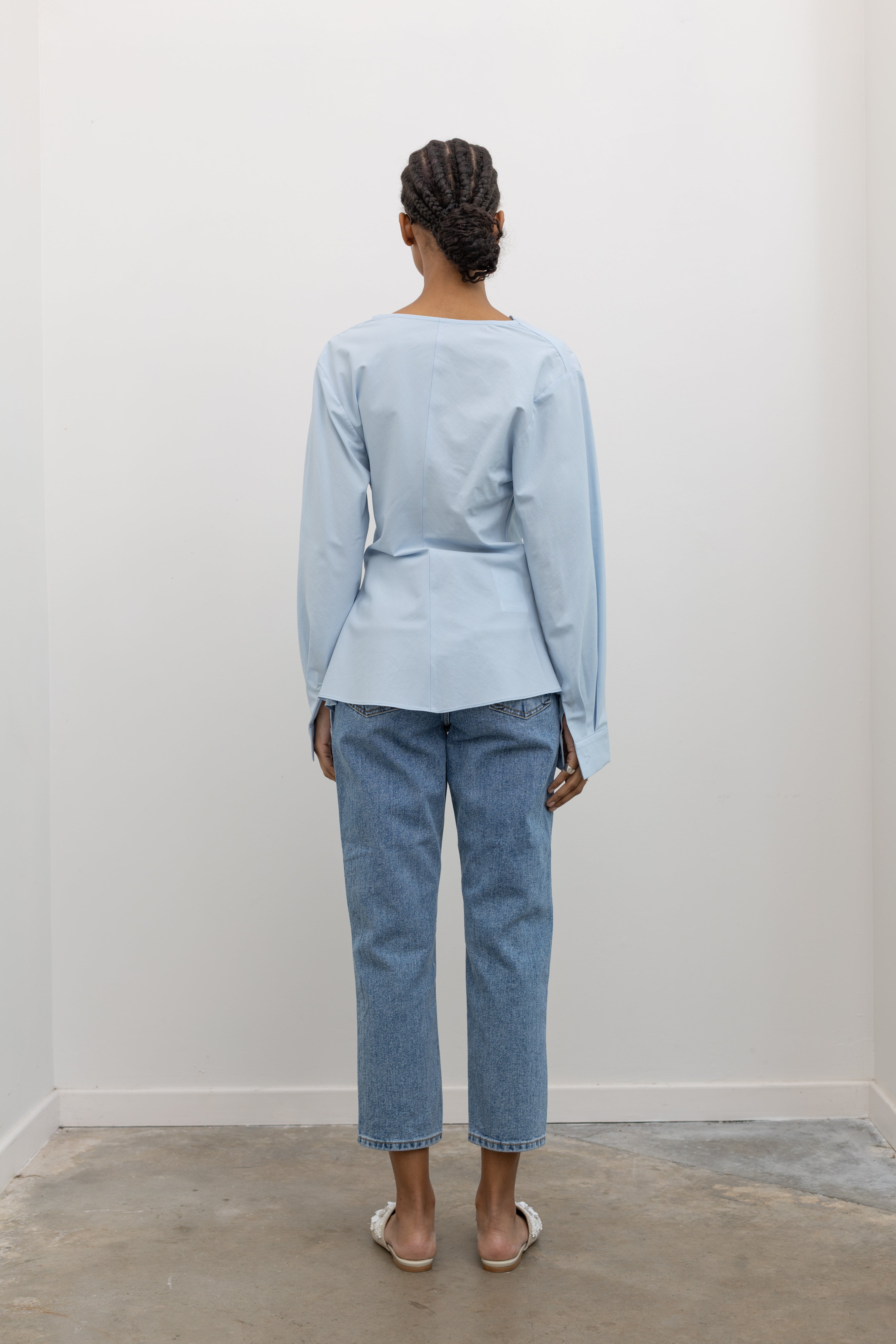 Full length back view of a woman wearing chic and architectural spring fashion in a Collective label sky blue cotton long sleeve top featuring a fitted natural waistline with a side draped motif creating an asymmetrical hem and cowl neckline paired with light wash denim jeans at Alas Eius. 