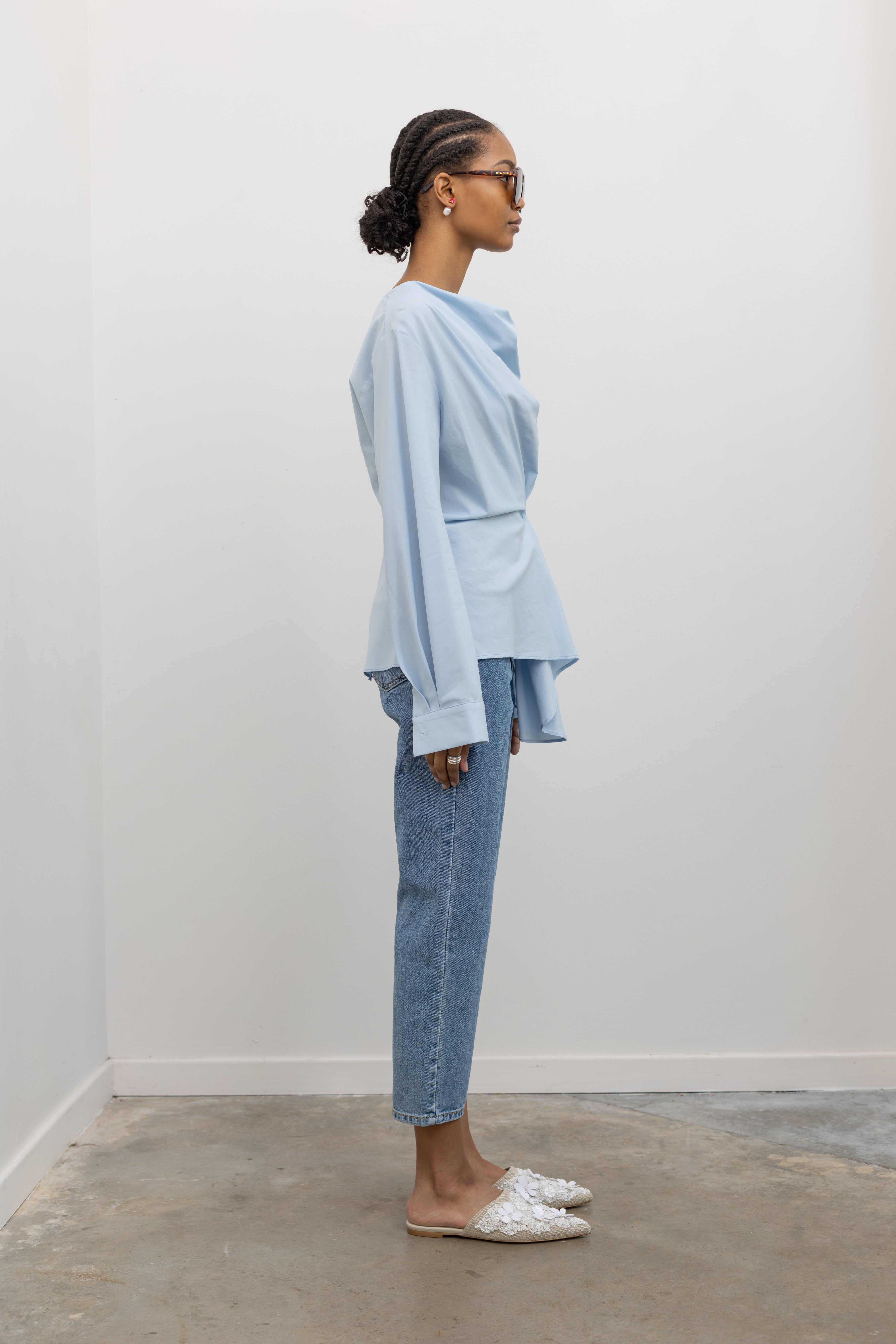 Full length side view of a woman wearing chic and architectural spring fashion in a Collective label sky blue cotton long sleeve top featuring a fitted natural waistline with a side draped motif creating an asymmetrical hem and cowl neckline paired with light wash denim jeans at Alas Eius. 