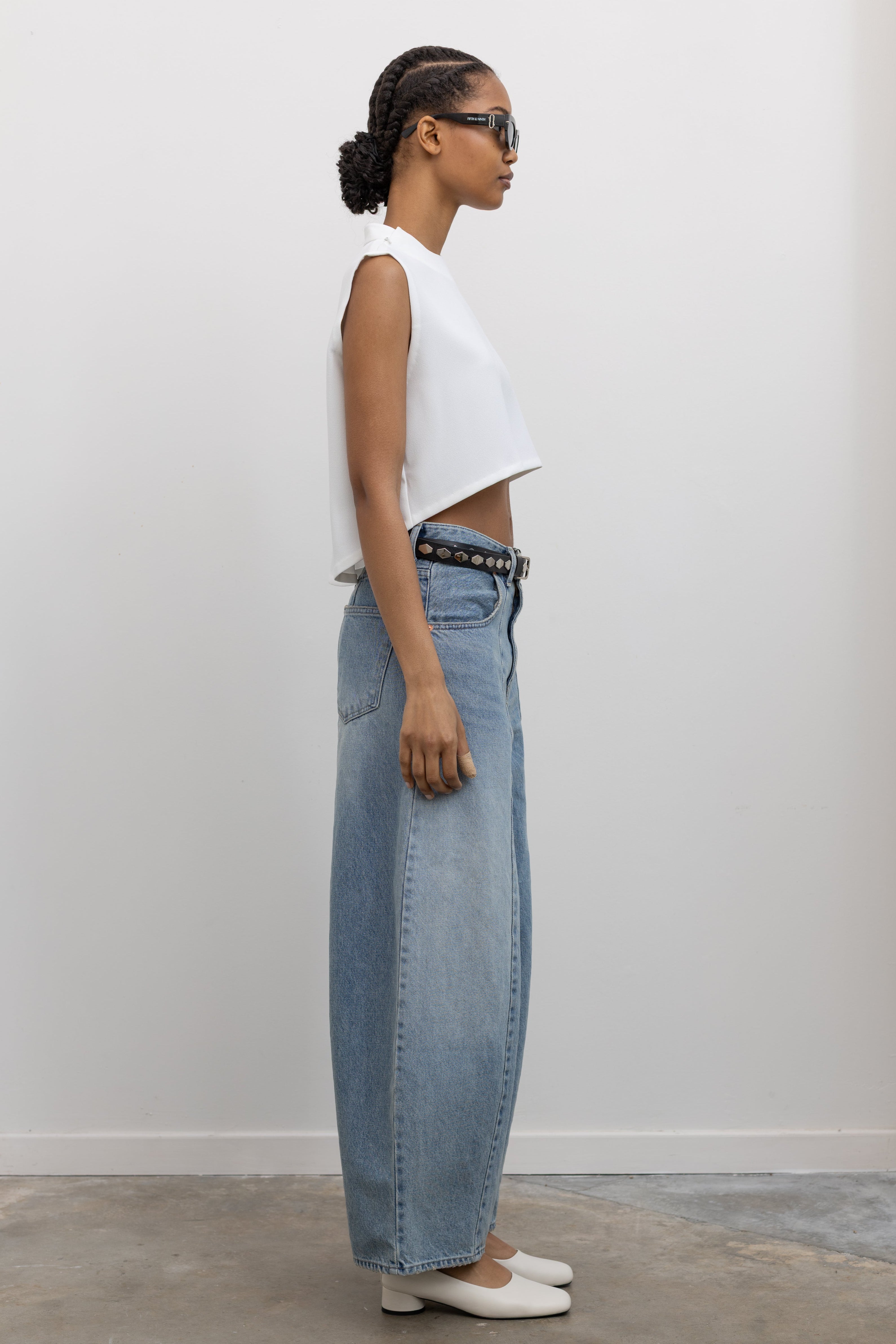 Side view of woman standing wearing Spring fashion in a white sleeveless cropped top and a pair of Collective Light Wash Front Seam Barrel Jeans with black leather studded belt and white pumps at Alas Eius