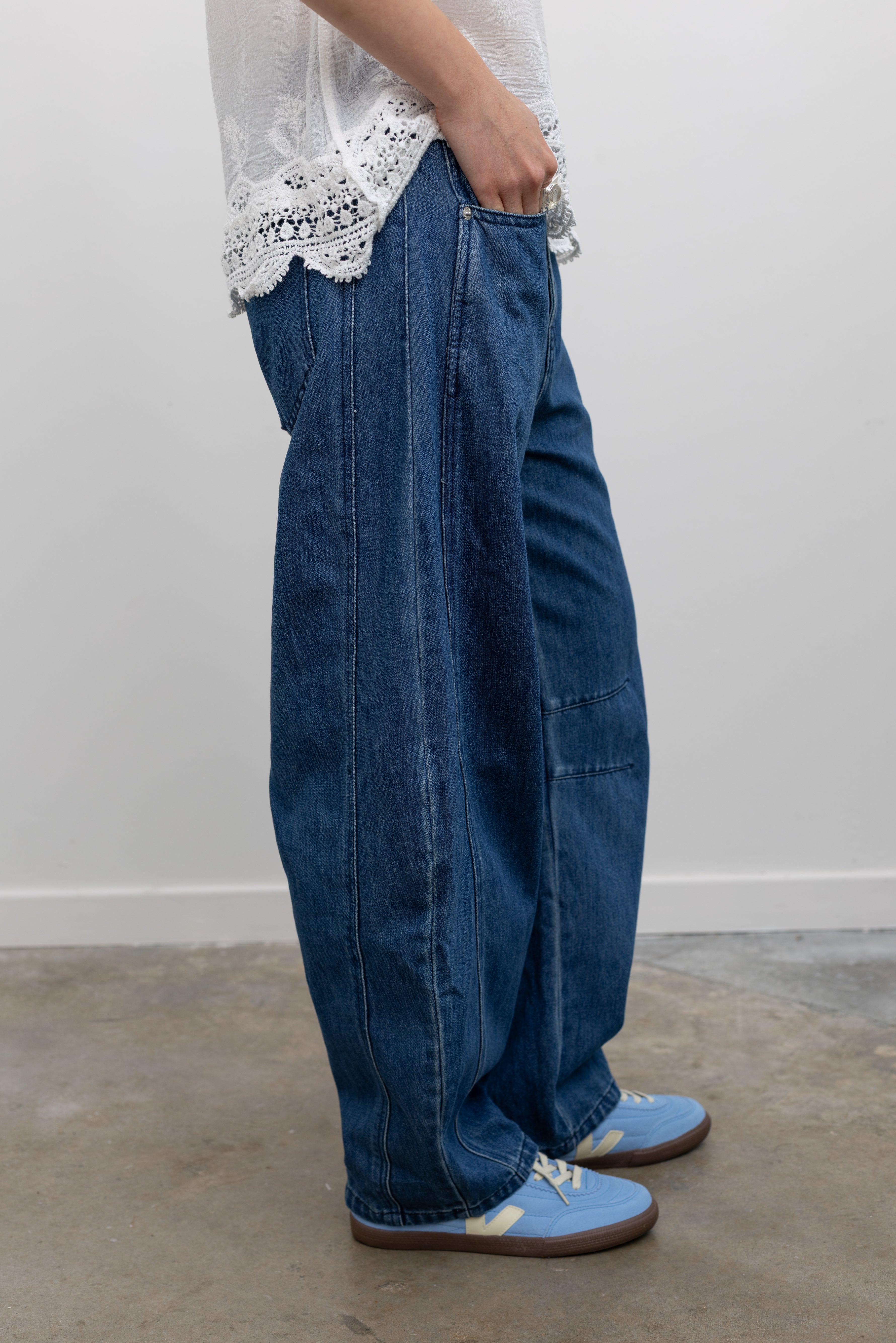 Model wearing effortless everyday jeans in the Collective blue cotton baggy denim pants with curved balloon silhouette, mid-rise waist, and full-length hem paired with a crochet lace camisole tank top and Veja blue suede sneakers at Alas Eius. 