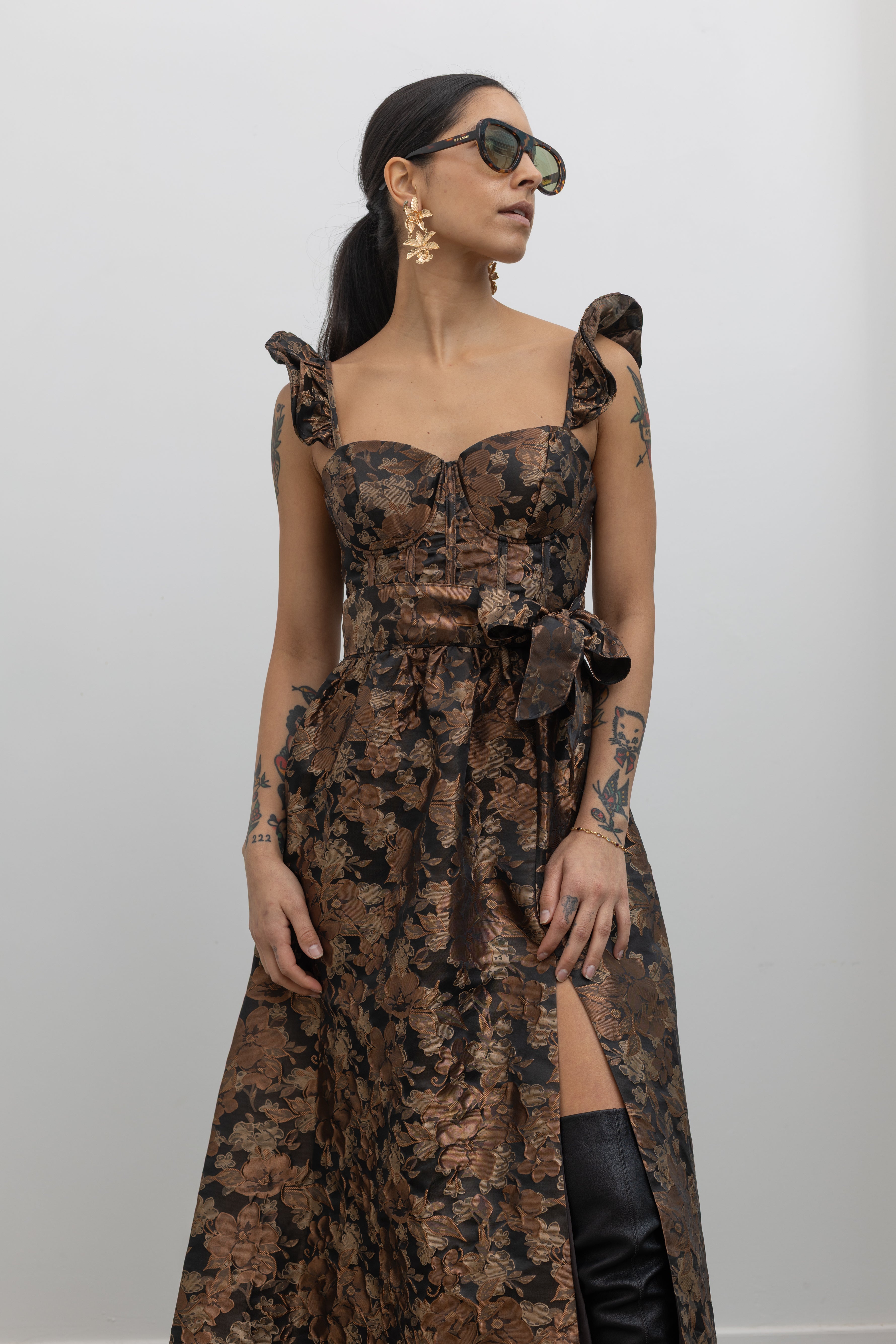 A woman posing wearing an elegant and feminine Collective label bronze floral jacquard corset midi dress featuring a high front slit and paired with over-the-knee slouchy black leather heeled boots at Alas Eius.