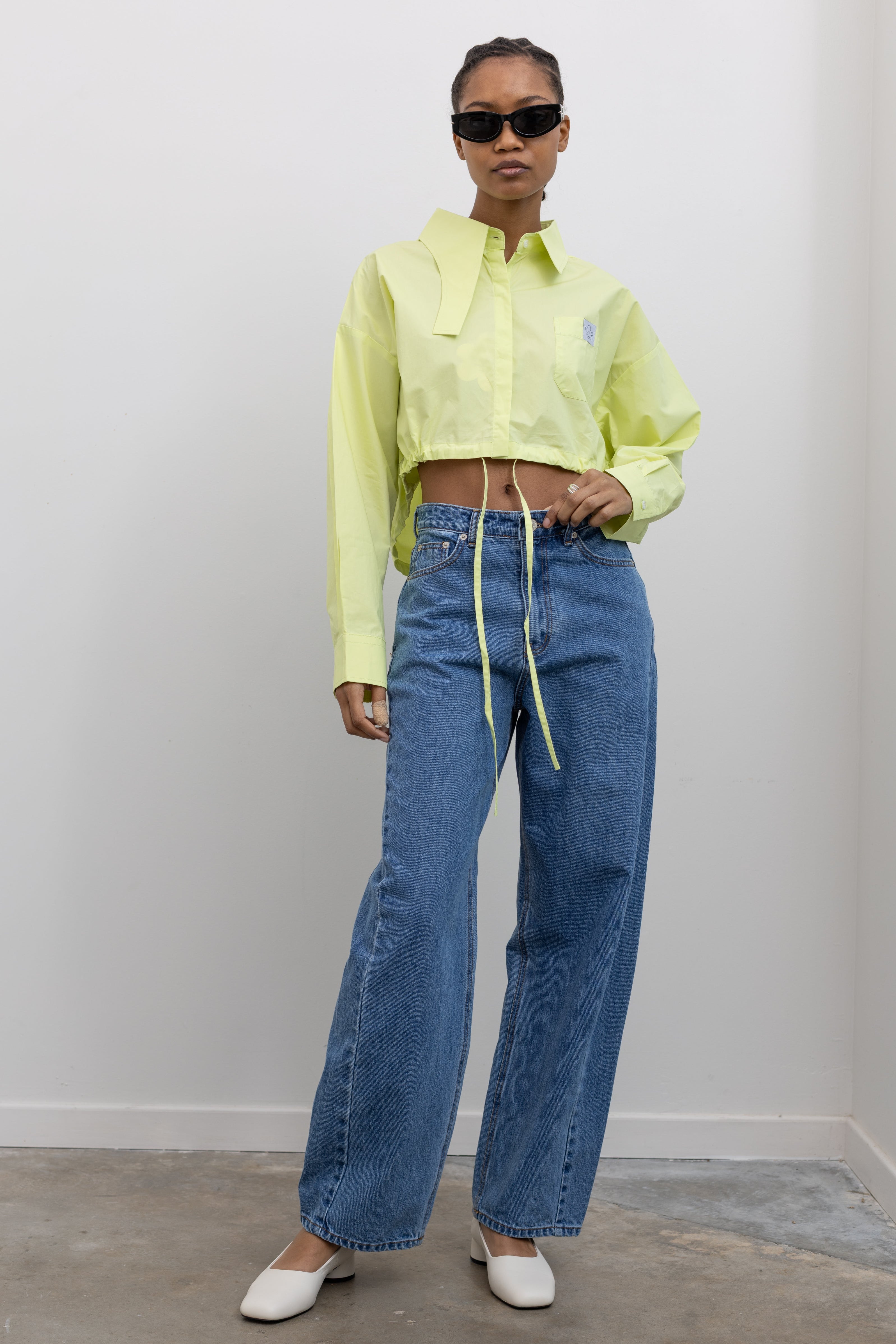 Woman standing wearing Spring fashion in a pair of Collective Mid Rise Baggy Balloon Stone Washed Jeans and a yellow cropped shirt with sunglasses at Alas Eius