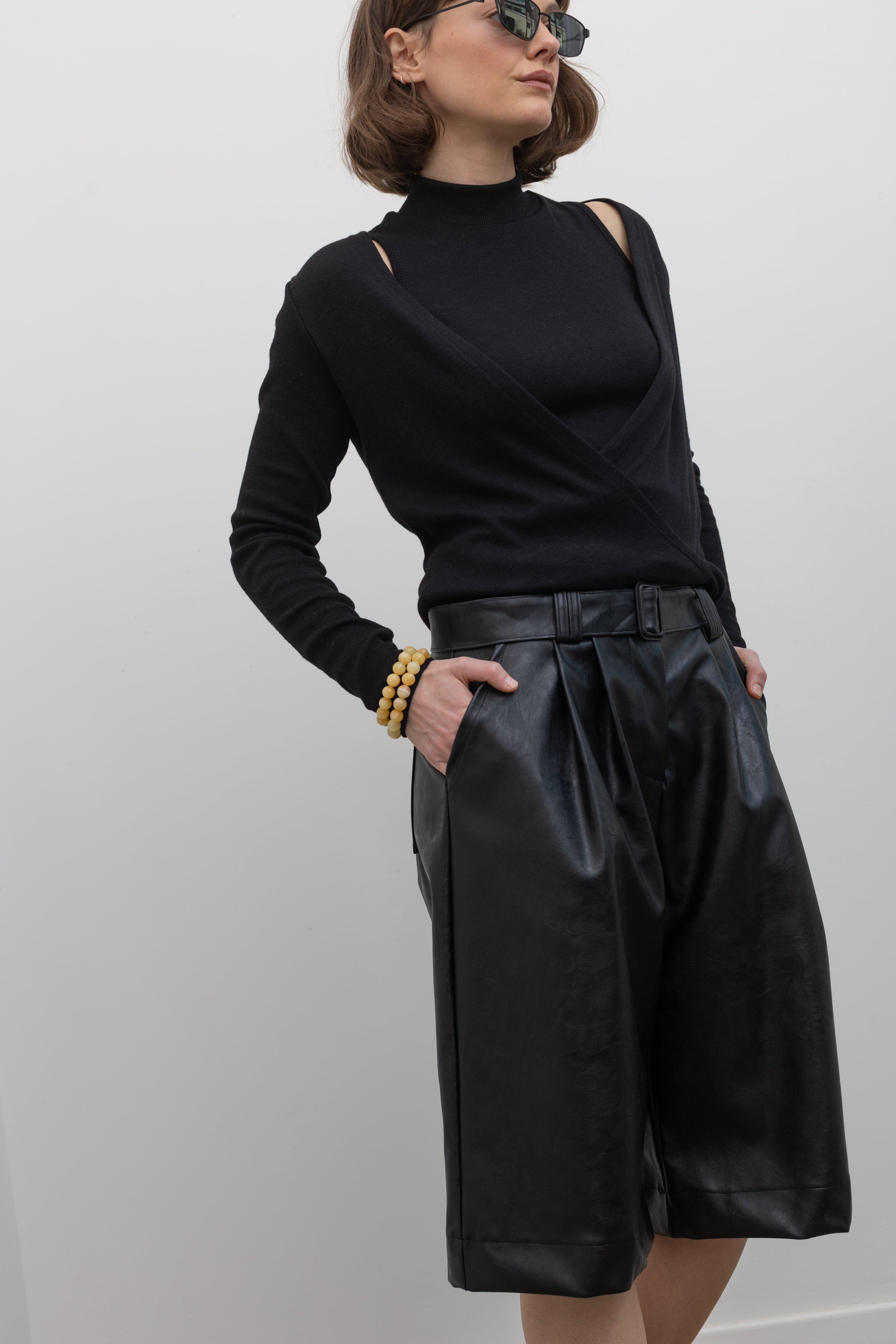 Close-up 3/4 view of a woman wearing Collective label Black Vegan Leather Belted Bermuda Shorts with an Alas Eius label mock neck sweater and slingback kitten heels at Alas Eius.