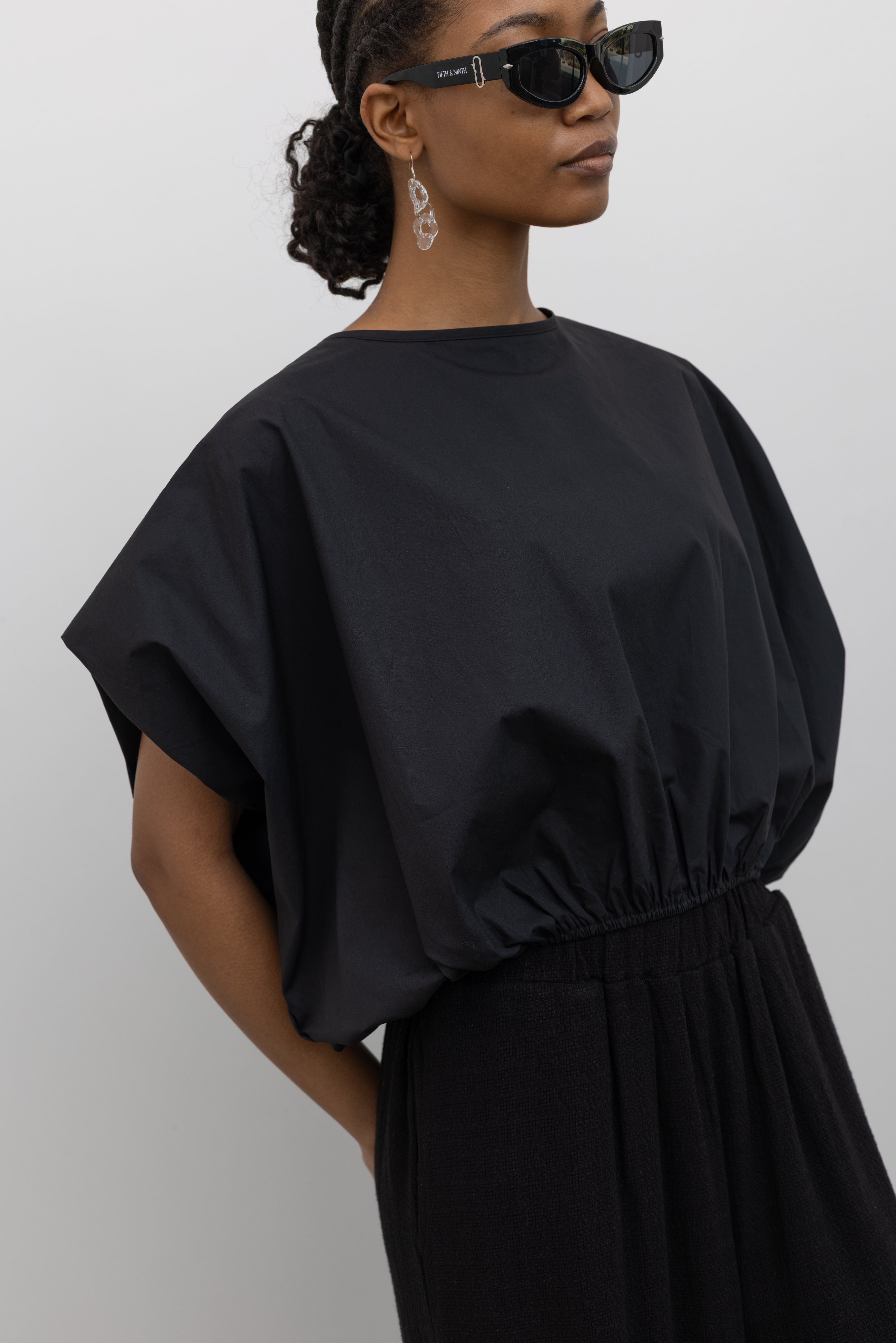 Close-up front view of a woman wearing Spring fashion in a contemporary design Collective Black Cotton billow cinched top featuring draped rounded sleeves, an elasticized waistband for a fitted torso and ribbon tie at the back neck at Alas Eius. 