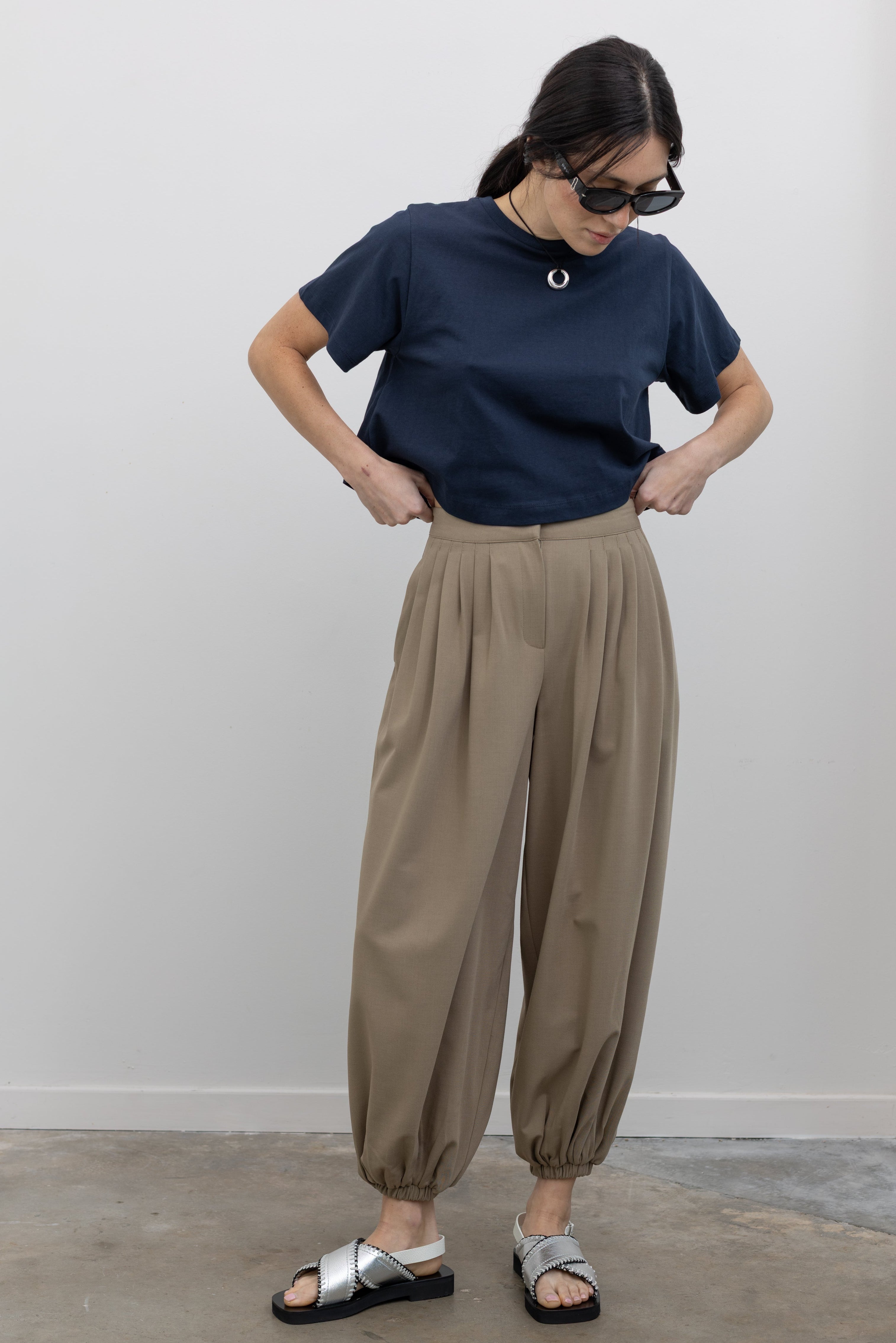 Full length front view of a woman wearing modern minimalist spring fashion in a Collective brand beige Tores pleated front pant featuring a cinched elastic ankle creating a balloon silhouette paired with an Alas Eius label navy blue t-shirt and metallic silver sandals at an Alas Eius store.
