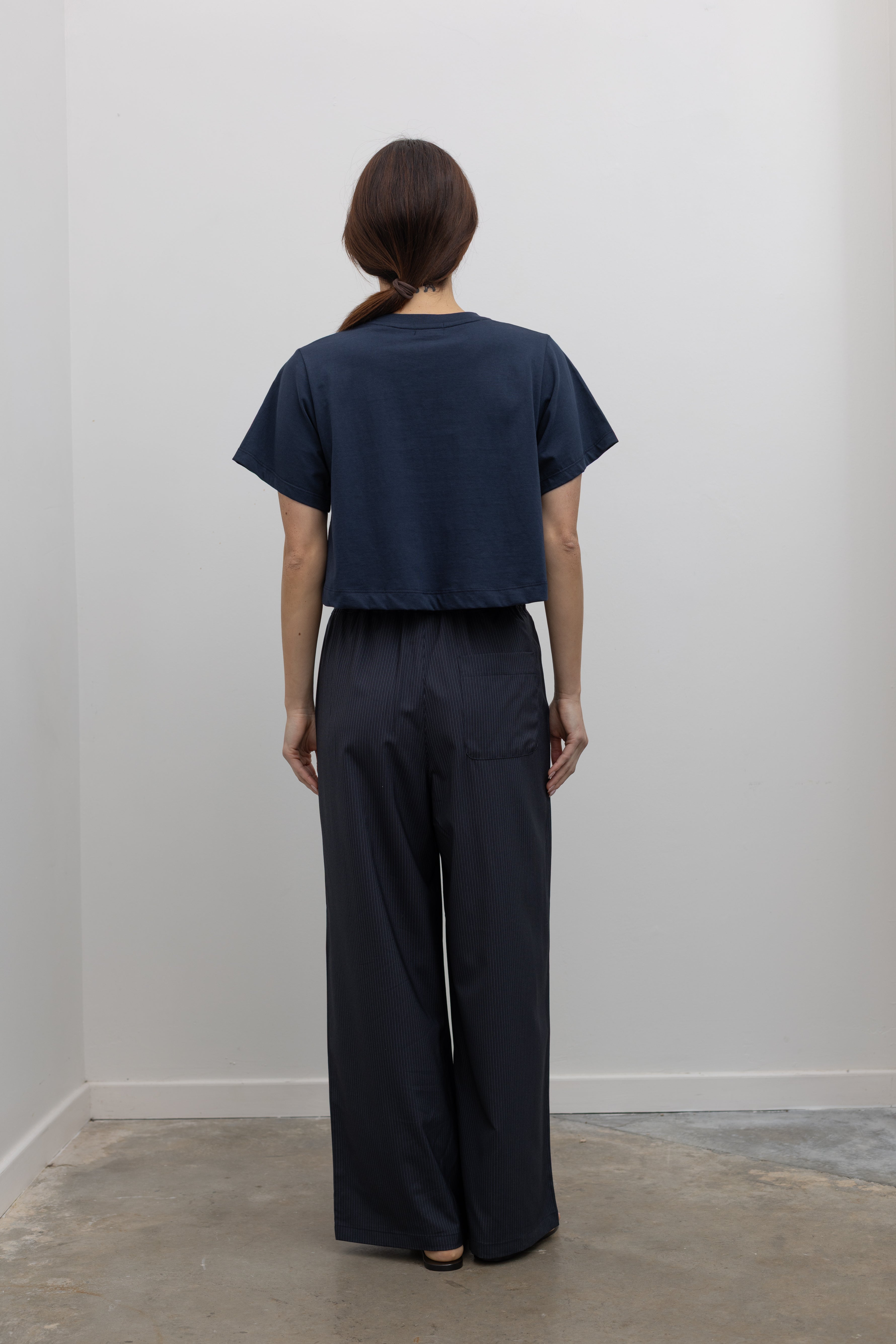 Back of Woman standing wearing Spring fashion in a pair of Collective Navy Pinstripe Wide Leg Drawstring Pants and navy blue tee with black leather sandals at Alas Eius