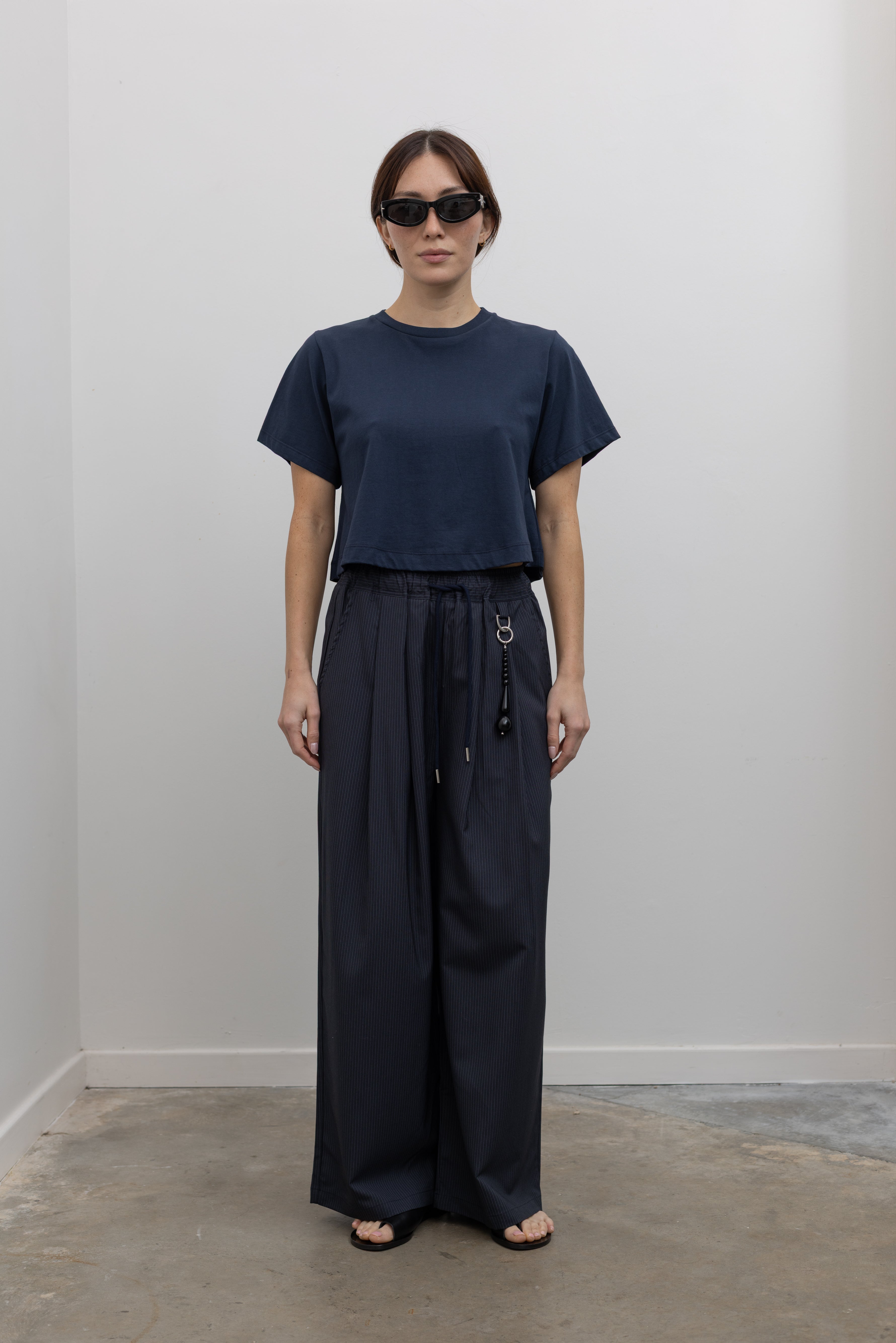 Woman standing wearing Spring fashion in a pair of Collective Navy Pinstripe Wide Leg Drawstring Pants and navy blue tee with black leather sandals at Alas Eius