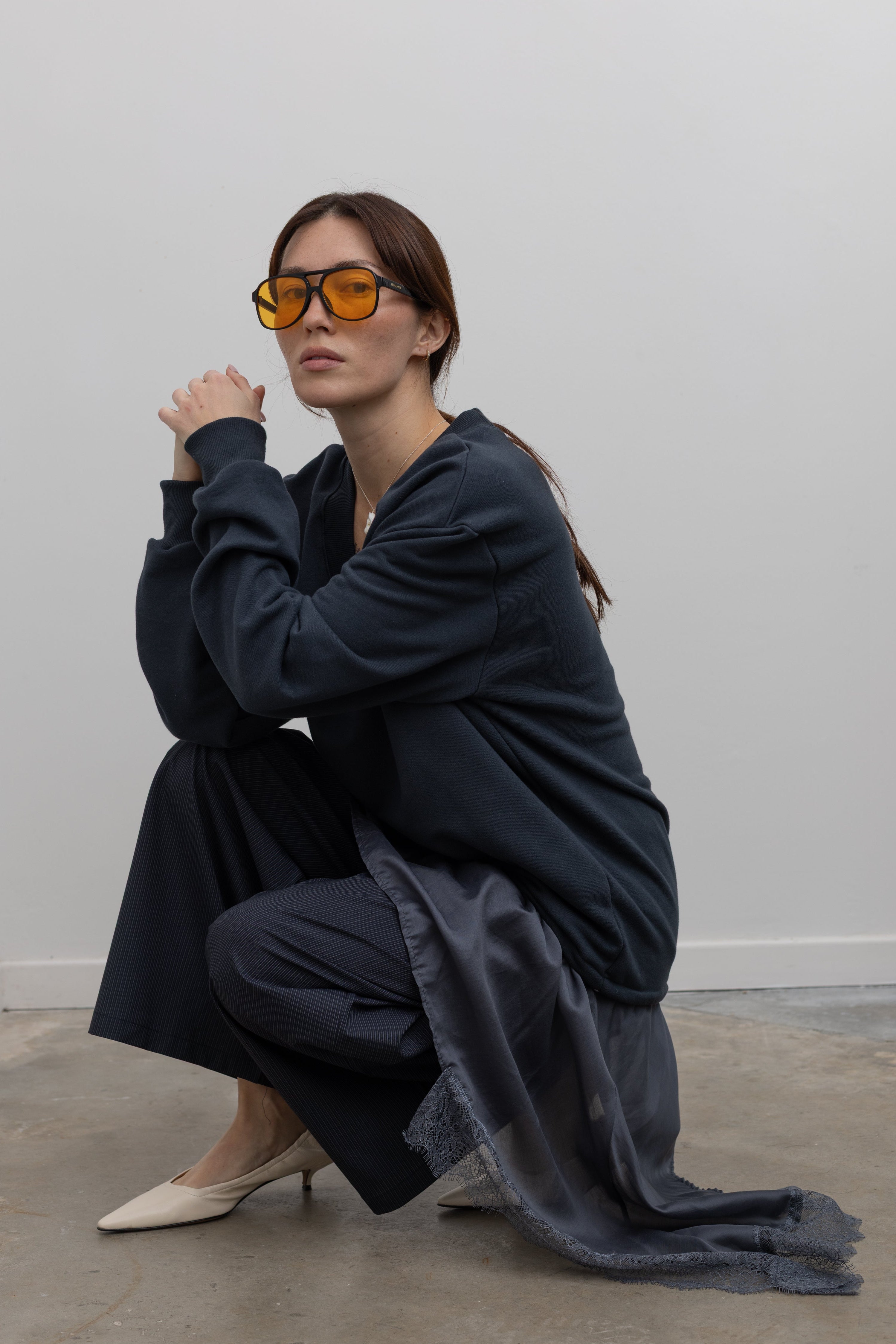 Woman kneeling wearing Spring fashion in a Collective Navy Lace Trim Cotton Oversized Jumper and navy blue pinstripe trousers with sunglasses and white pumps at Alas Eius 
