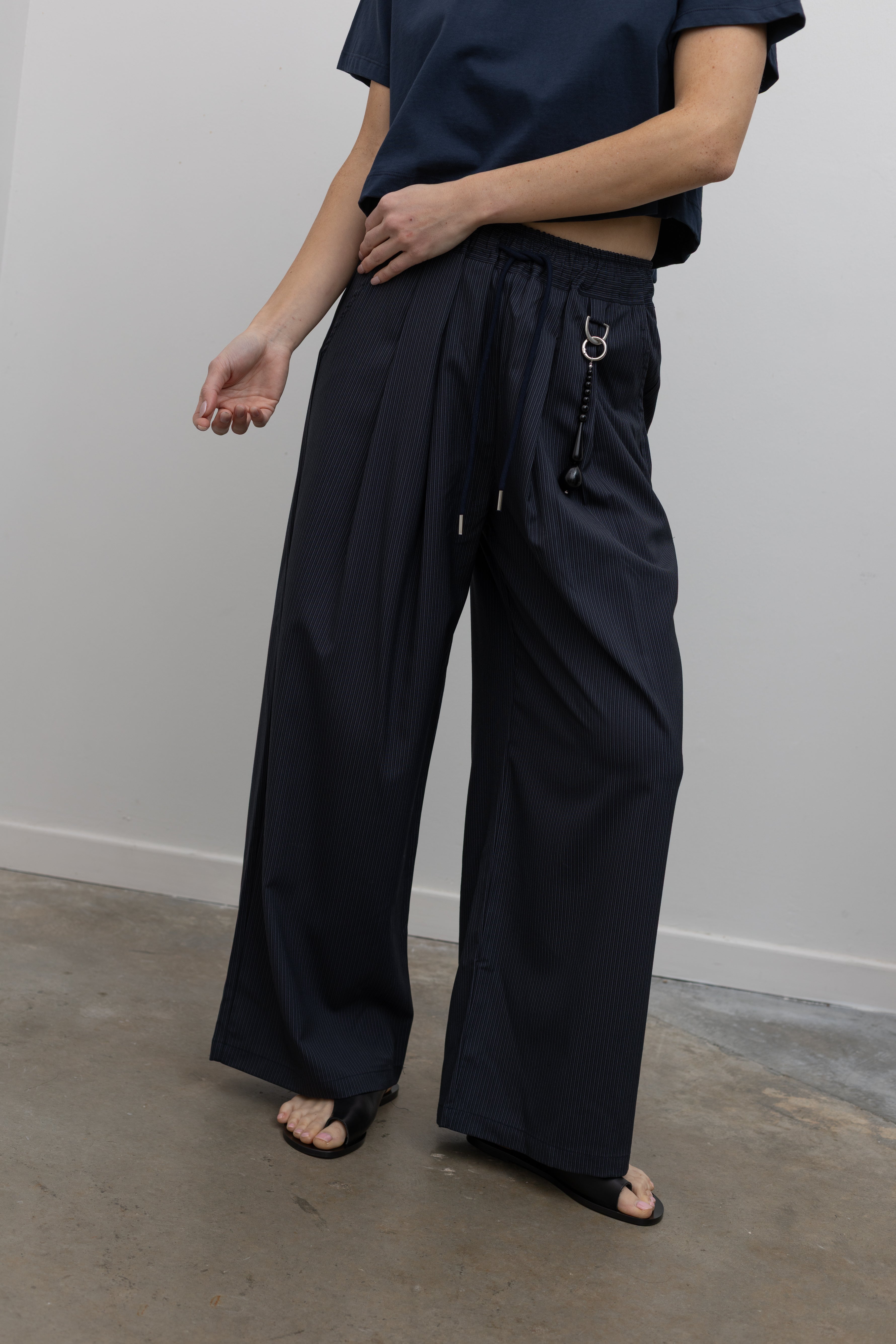 Woman standing wearing Spring fashion in a pair of Collective Navy Pinstripe Wide Leg Drawstring Pants and navy blue tee with black leather sandals at Alas Eius