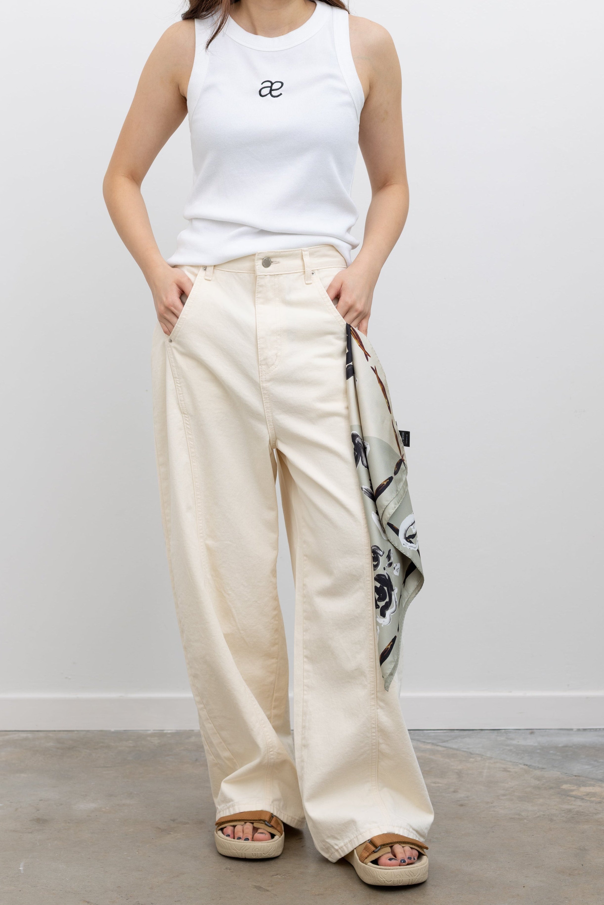 Model wearing elevated summer fashion in Collective off white heavy-weight cotton denim jeans with a structured wide-leg silhouette, curved paneling, and relaxed oversized fit paired with an Alas Eius label logo tank and Veja slide sandals at Alas Eius.